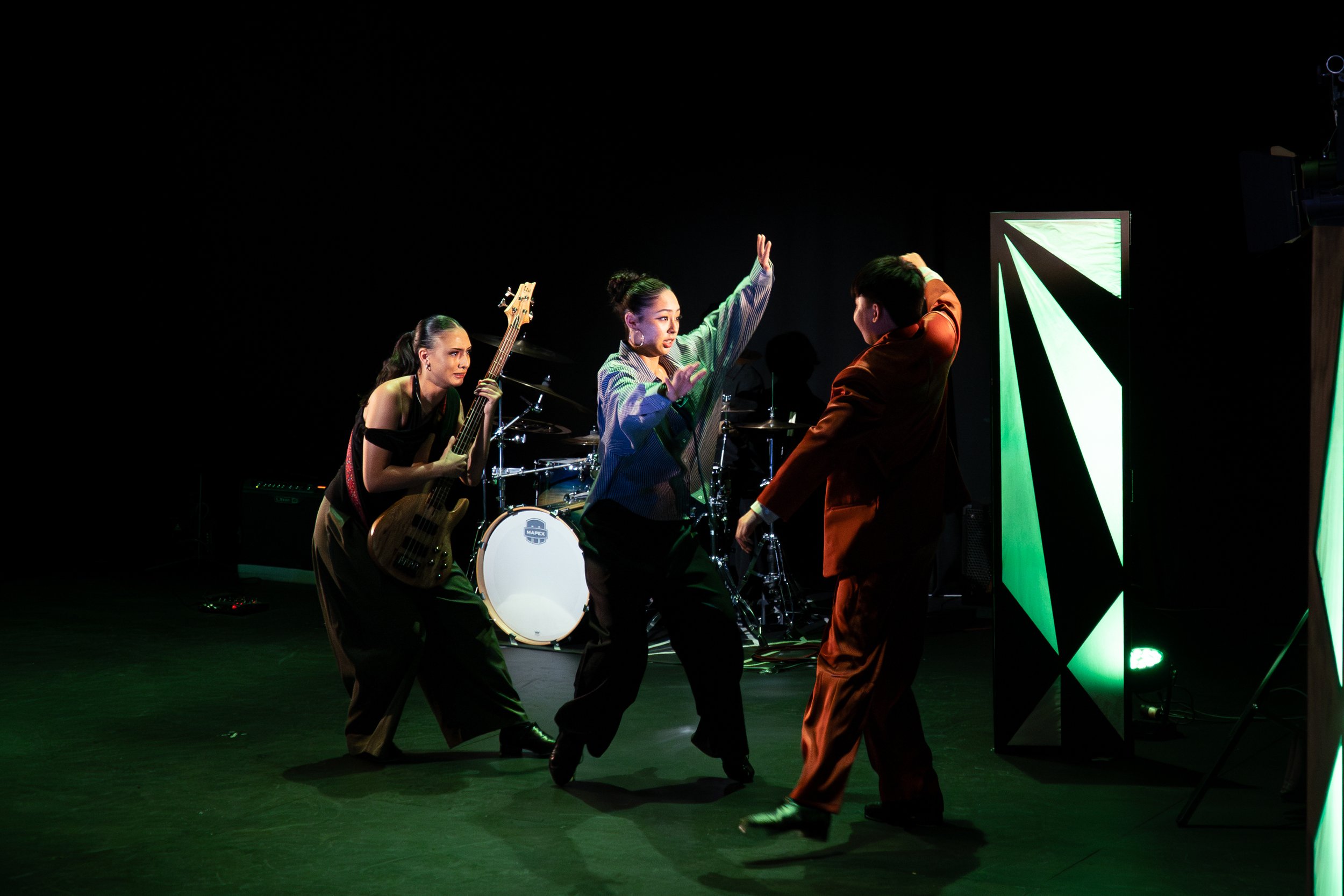 Four dancers performing with musical instruments and a decorative backdrop on stage. One woman plays a bass guitar, another is at a drum set, and two others are dancing.
