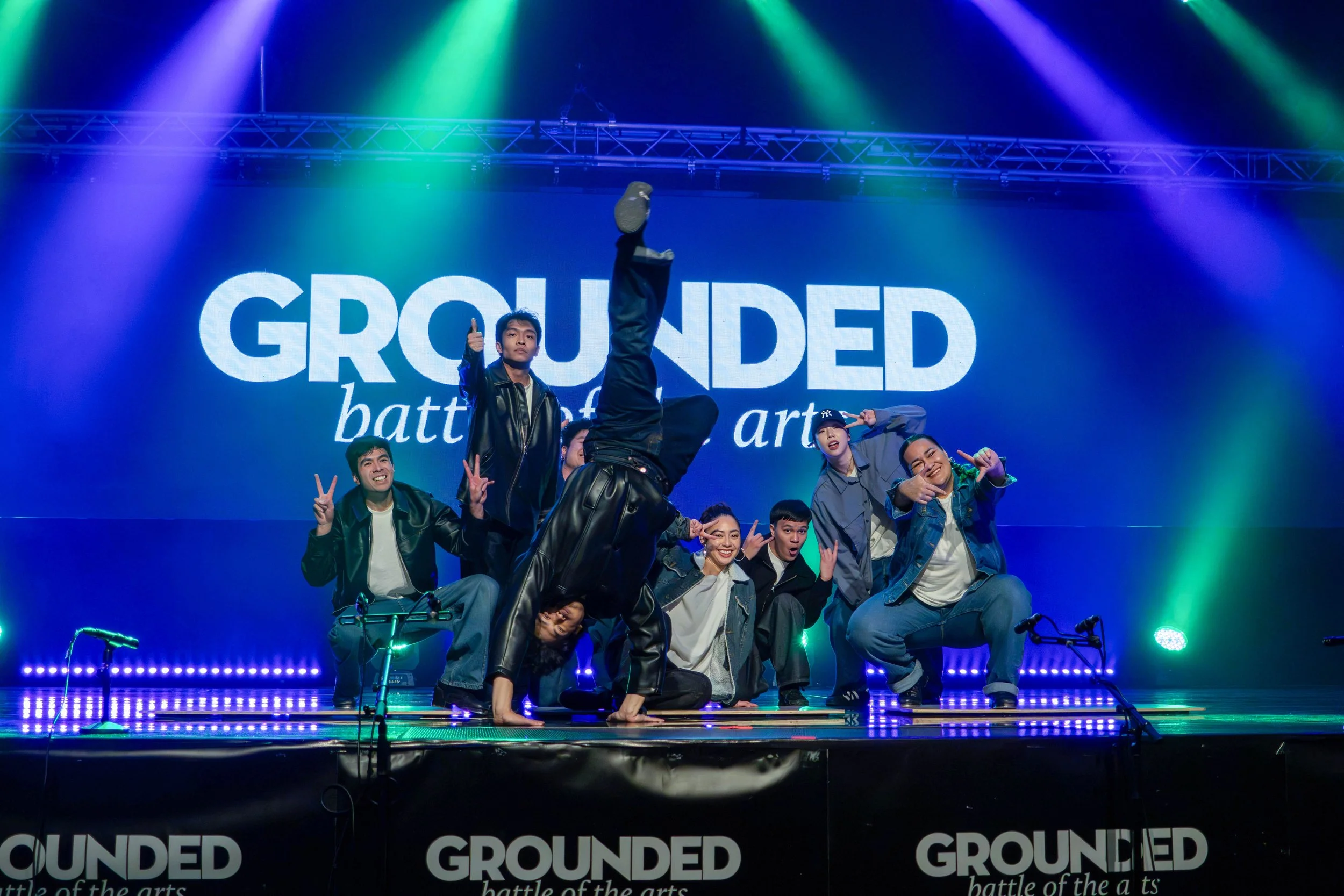 A group of dancers performing on stage with a large screen behind them displaying the words 'GROUNDED' and more text. The stage is lit with colorful lights.