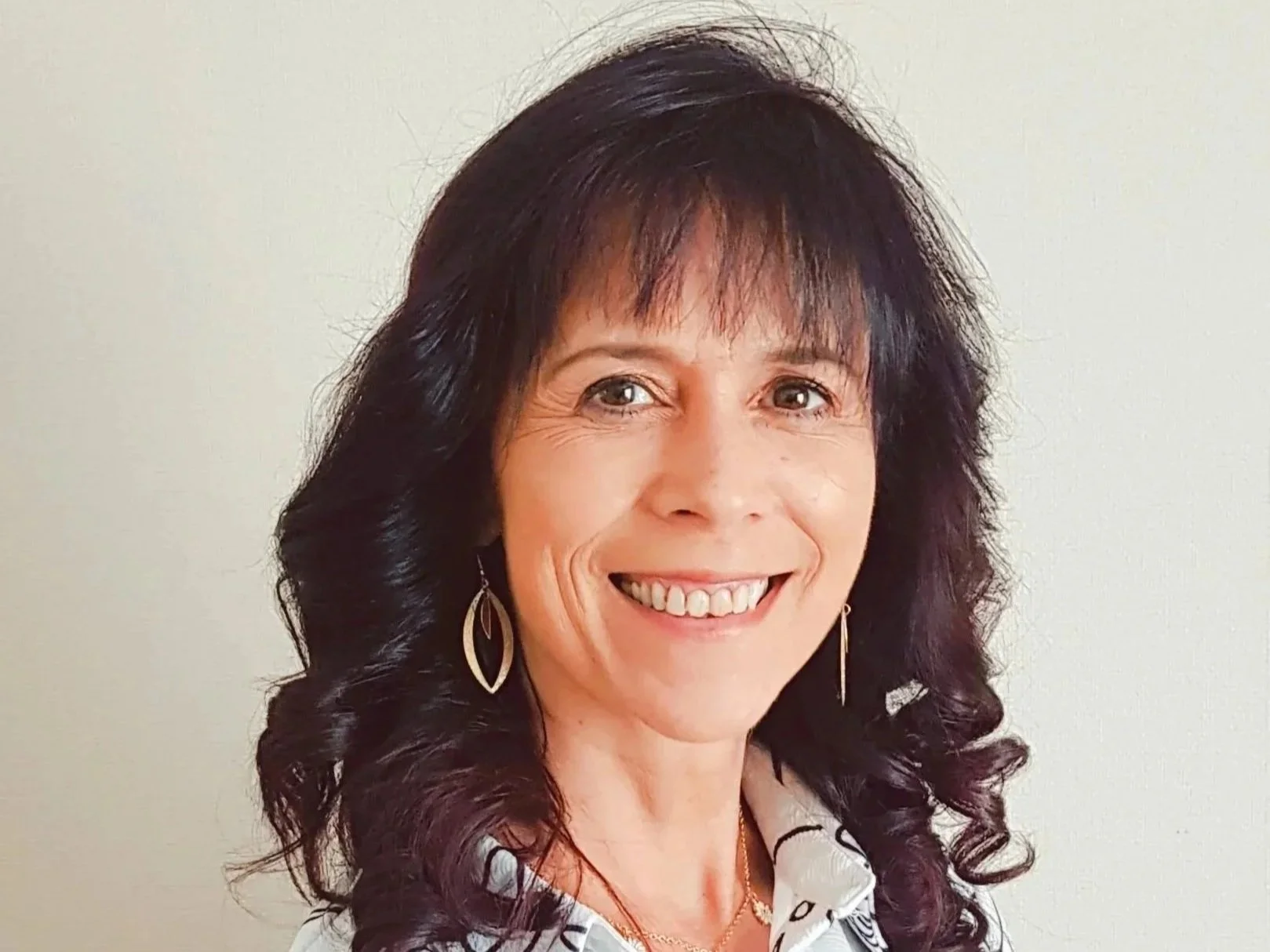 A woman with dark, wavy hair and light skin, smiling and wearing gold earrings and a white shirt with a pattern, standing against a plain light-colored wall.