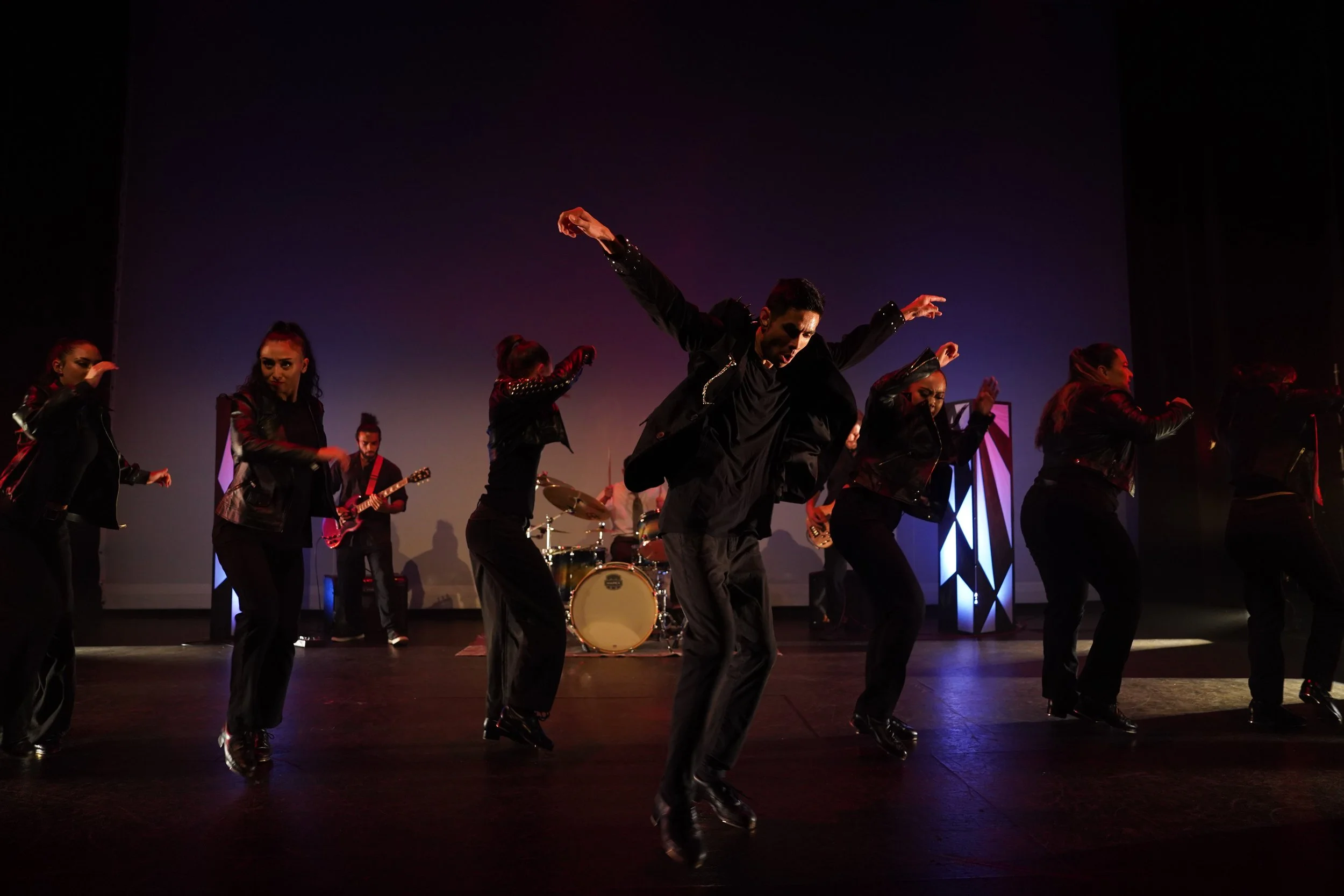 Dancers performing on stage with musical instruments in the background. The stage is dimly lit with purple and red lighting.