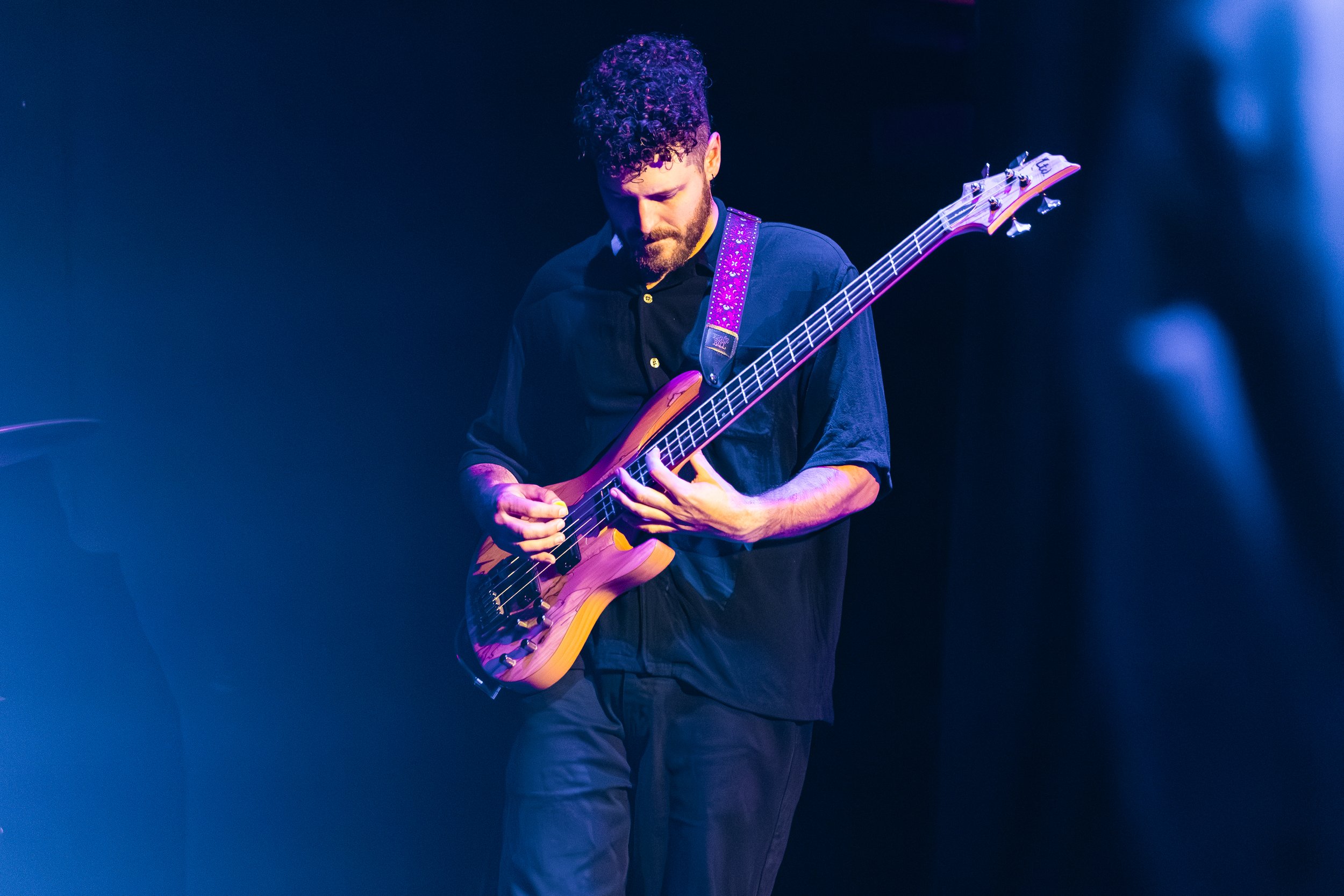 A man with curly hair and a beard is playing an electric guitar on stage, illuminated by blue and purple stage lights.