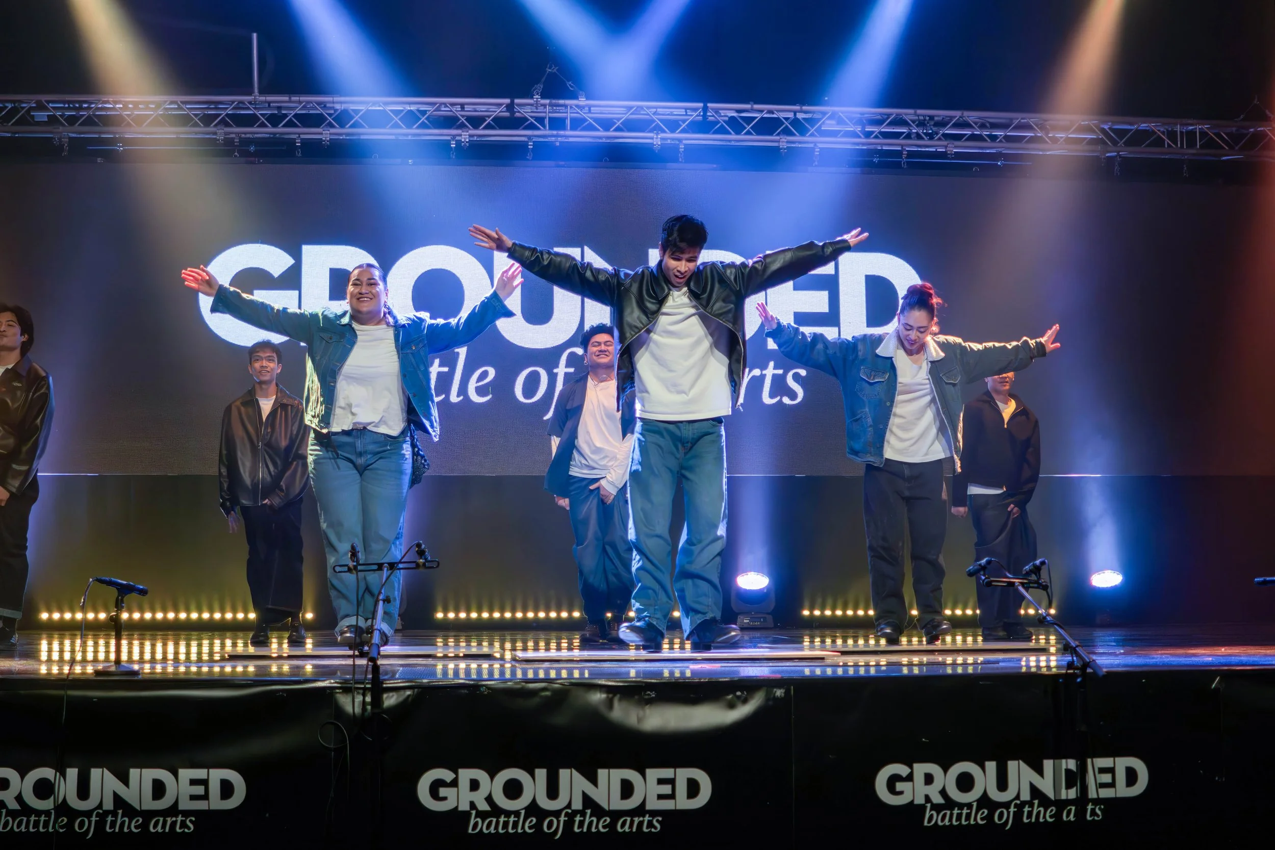 Group of dancers performing on stage with a large screen behind them displaying the word 'GROUNDED' and a tagline 'battle of the arts'. The stage is lit with colorful spotlights, and the performers are in casual clothing with arms outstretched.