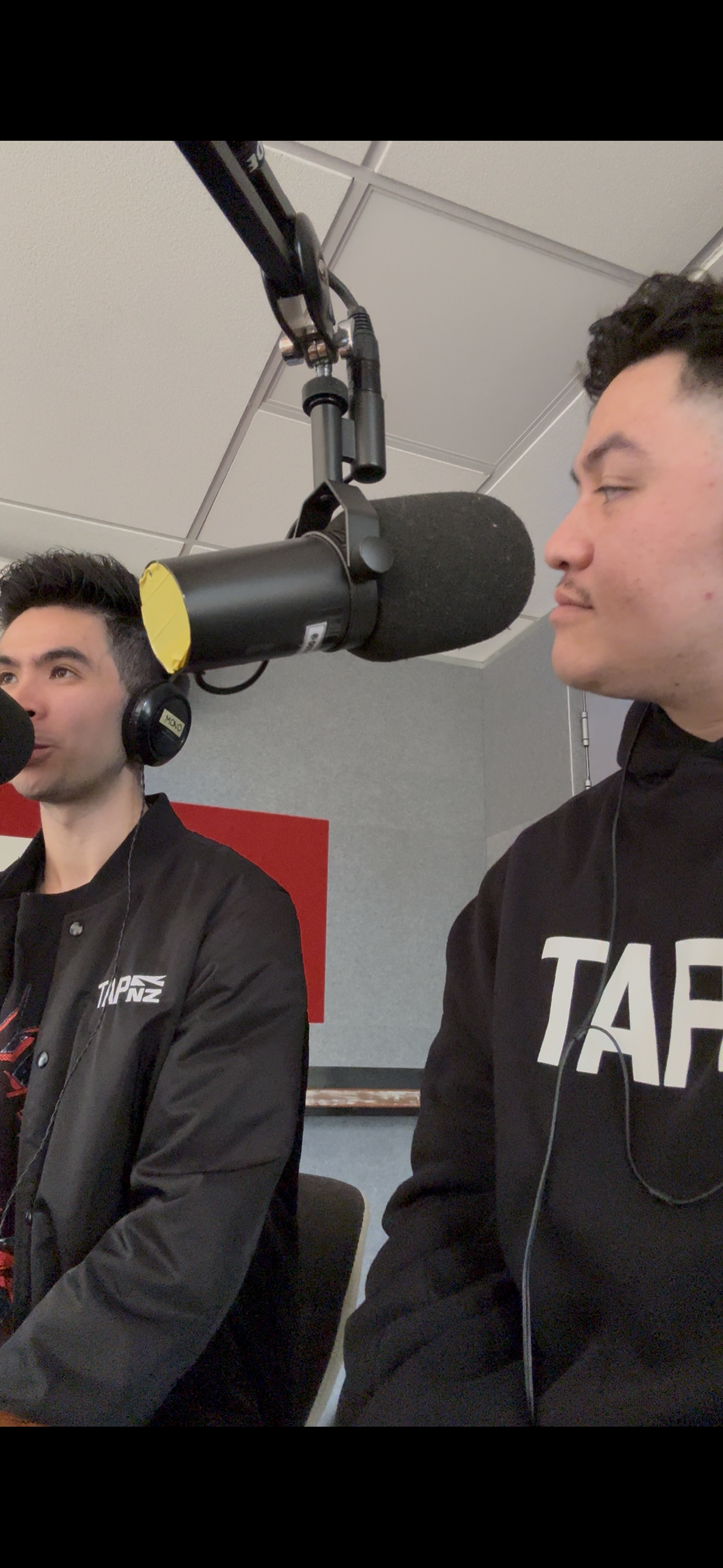 Two young men in a recording studio sit in front of microphones with closed eyes, wearing headphones and casual clothing, in a professional radio or podcast setting.