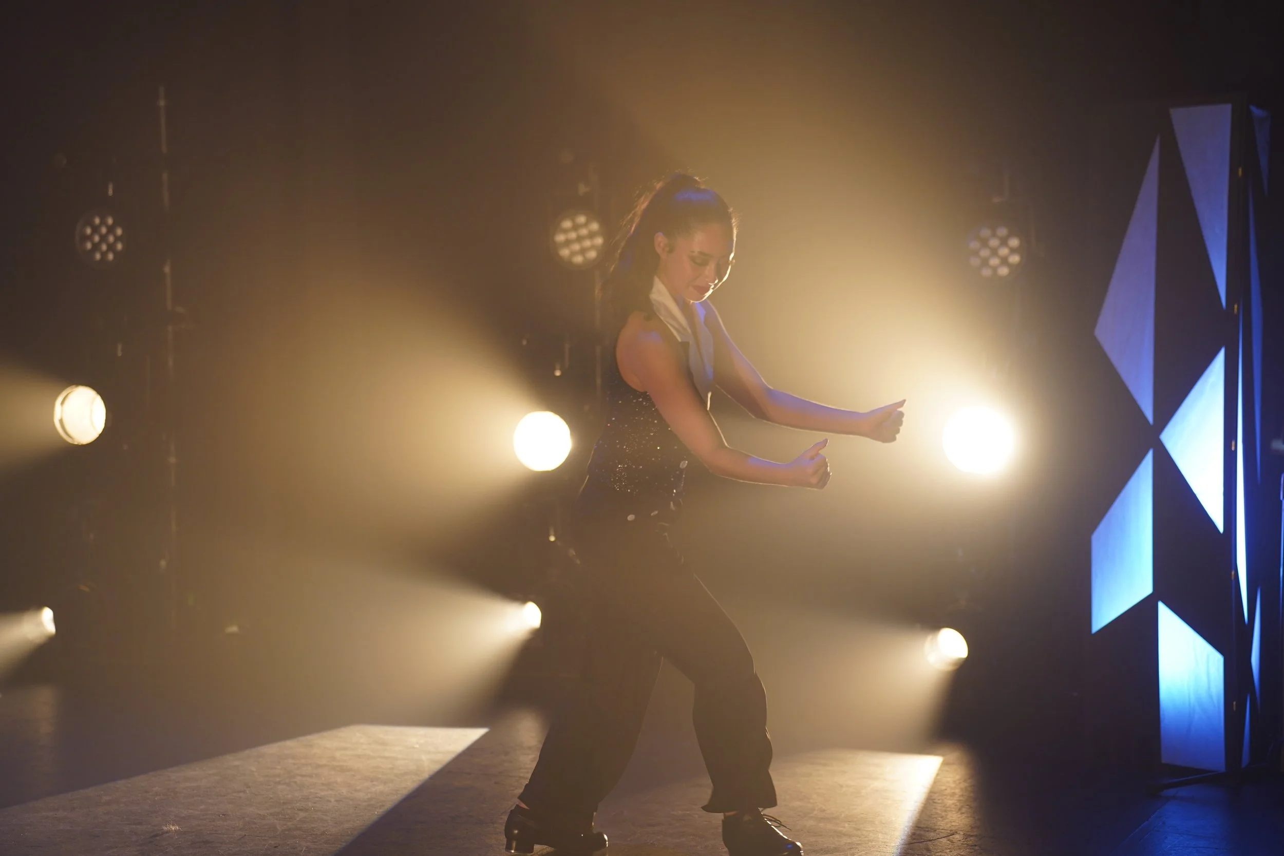 A woman dancing in a dark stage with bright backlighting and geometric blue light panels on the side.