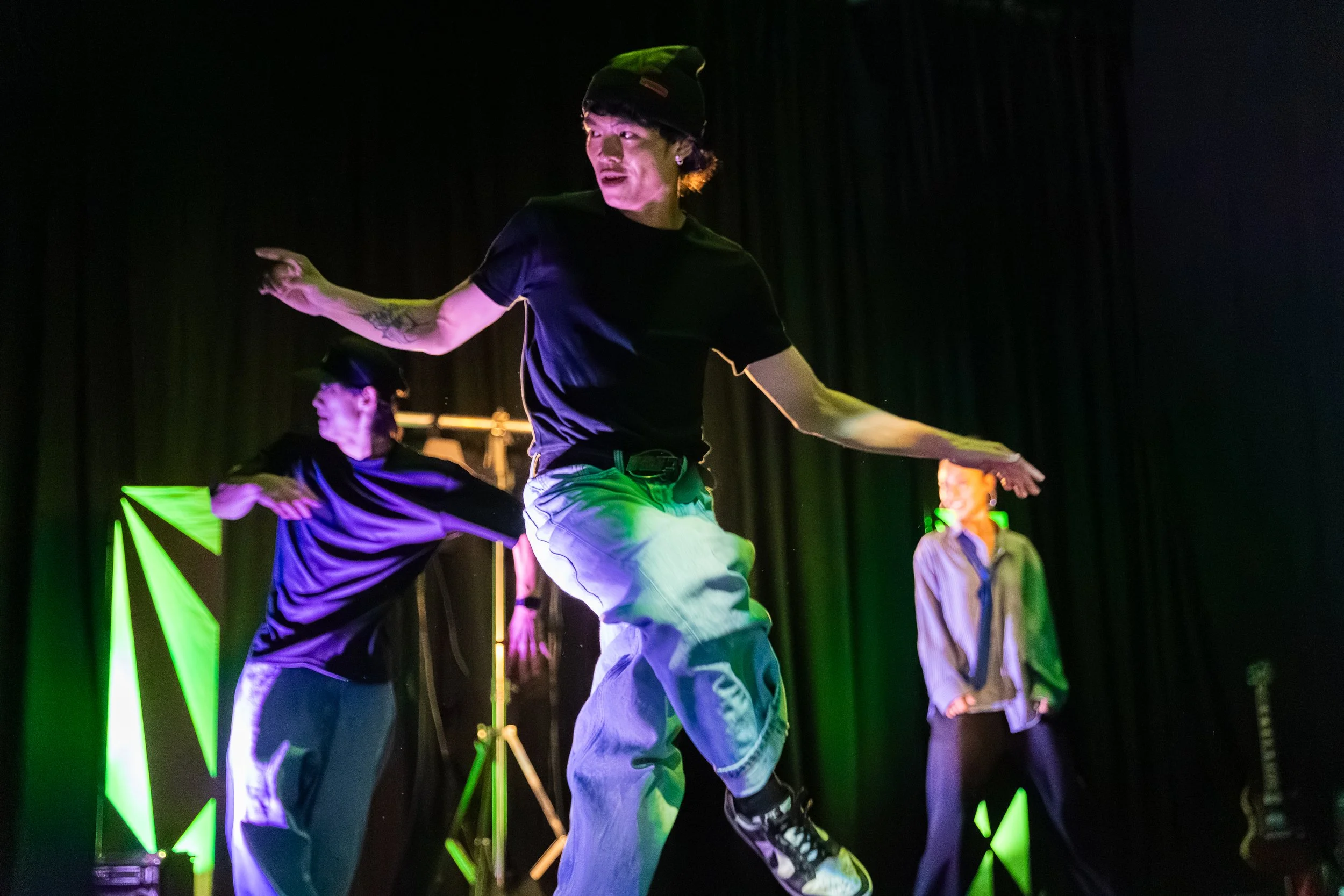 Performers dancing on stage with a dark background, wearing casual clothing including hats and sneakers, illuminated by colorful stage lights.
