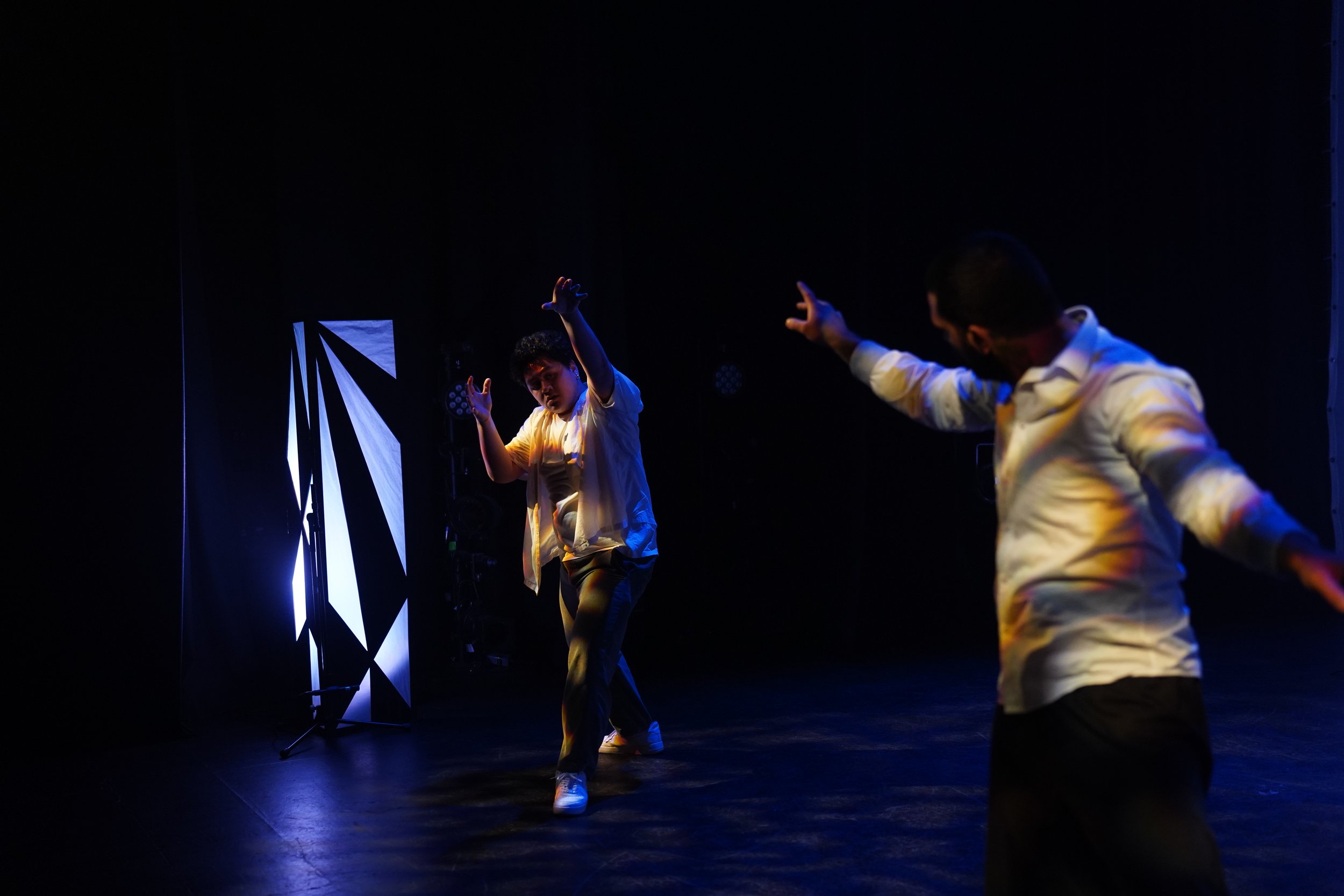 Two dancers performing on a dark stage with blue lighting, one dancer in the foreground with his hand raised and the other dancer in the background, both dressed in light-colored shirts and pants.