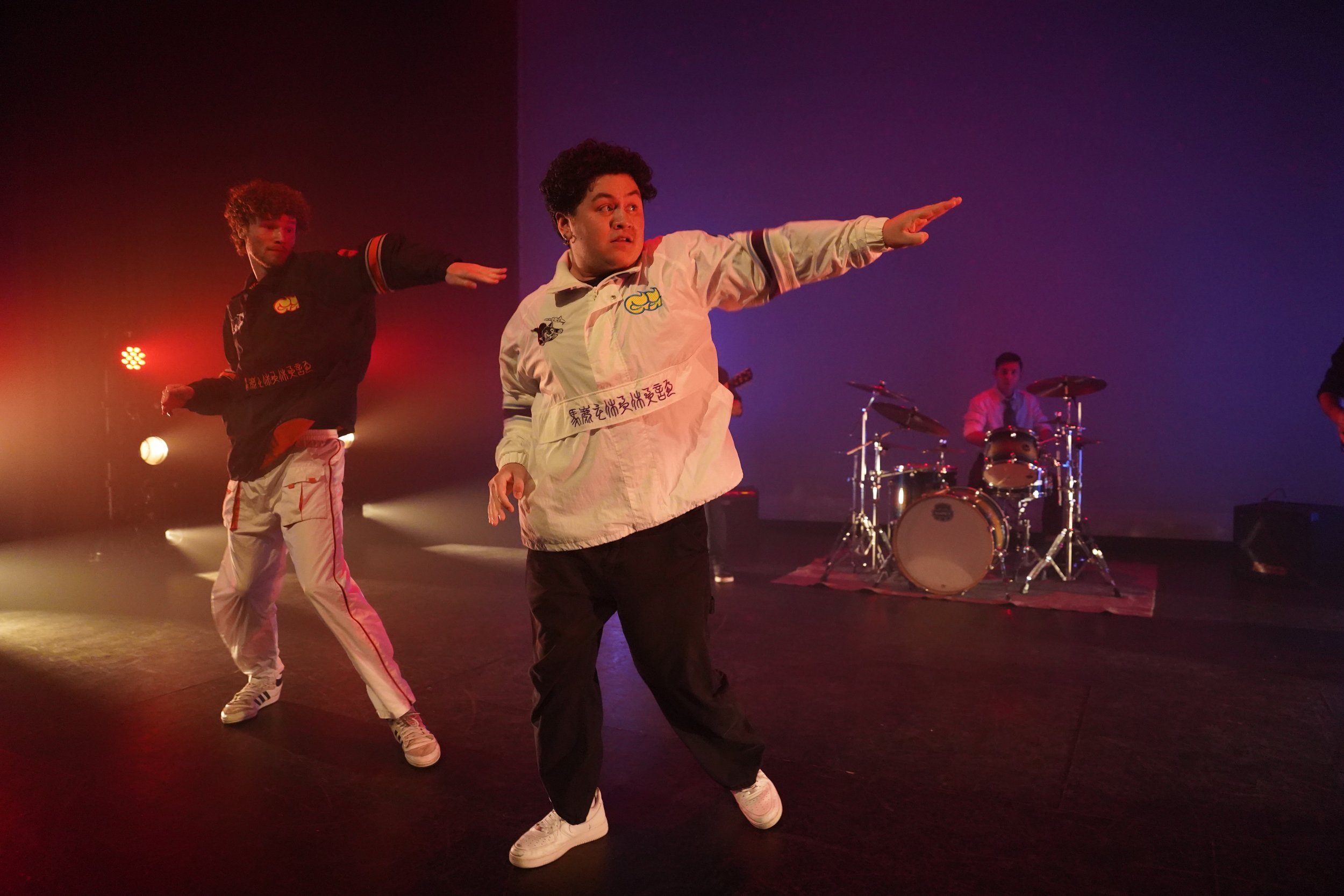 Two men dancing on stage with a drummer playing in the background, illuminated by colorful stage lighting.