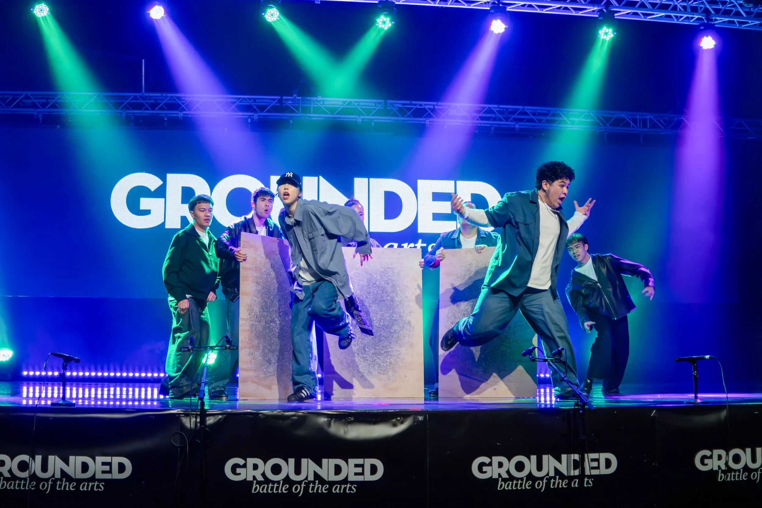 A group of performers dancing on stage with colorful lighting and a large screen behind them displaying the word 'GROUNDED'.