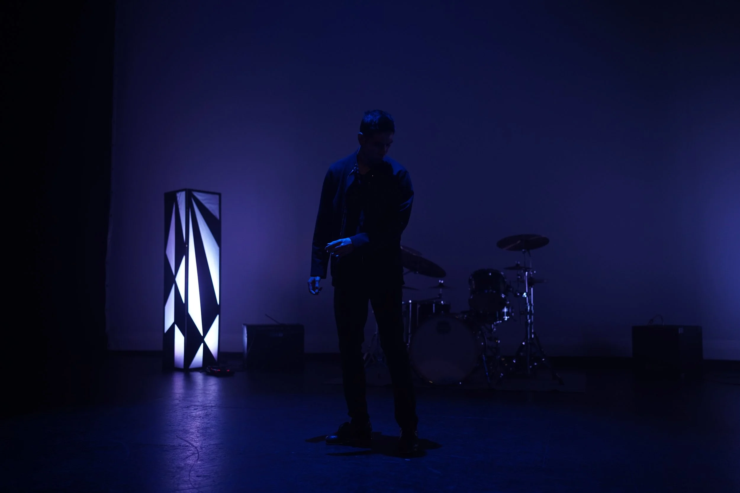 A person stands on a stage with dim blue lighting, surrounded by musical instruments, including drums, and illuminated by a tall, modern light fixture.