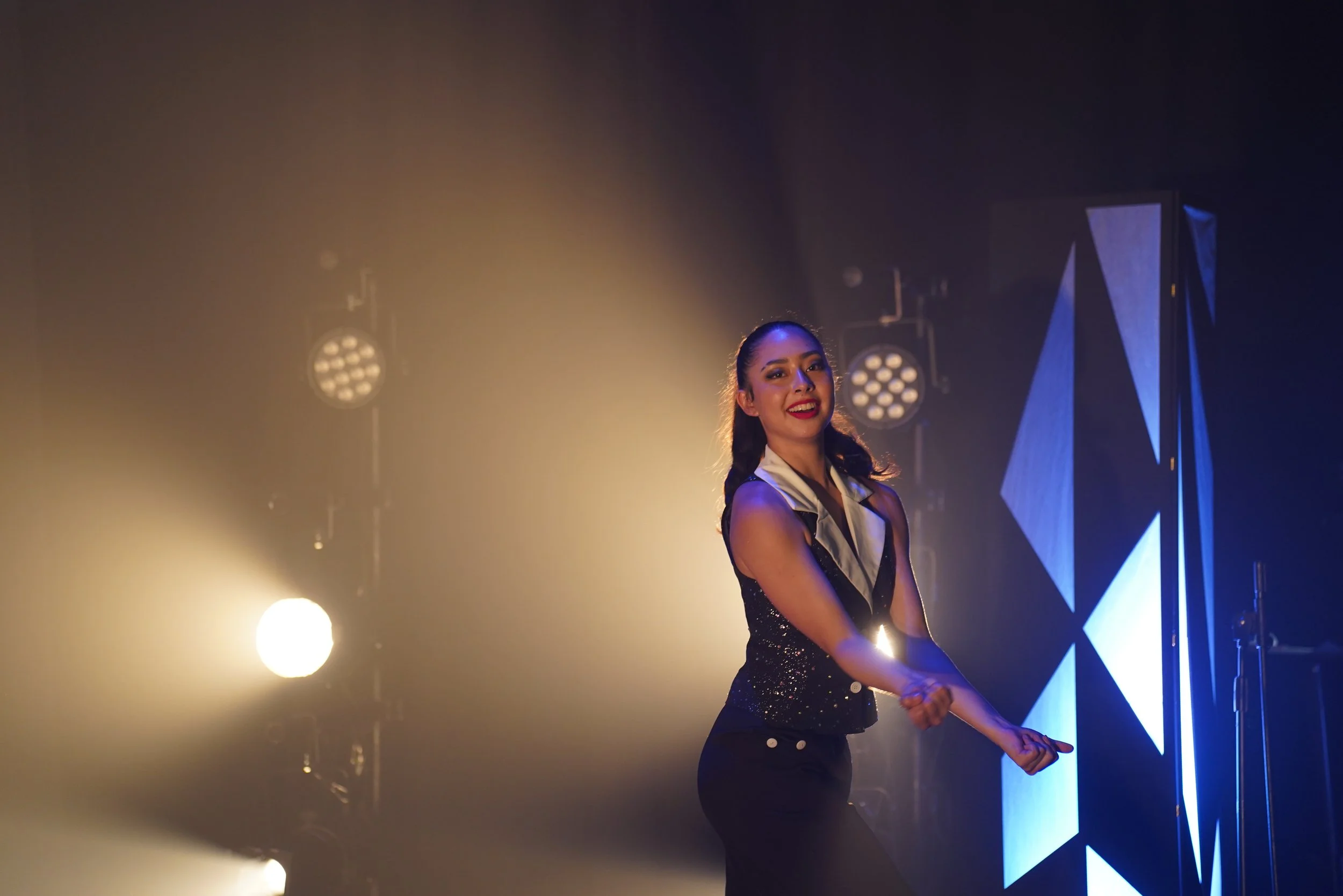 A woman with dark hair styled in waves, wearing a sparkly black dress and red lipstick, smiling and performing on stage with bright lights and geometric stage decorations in the background.