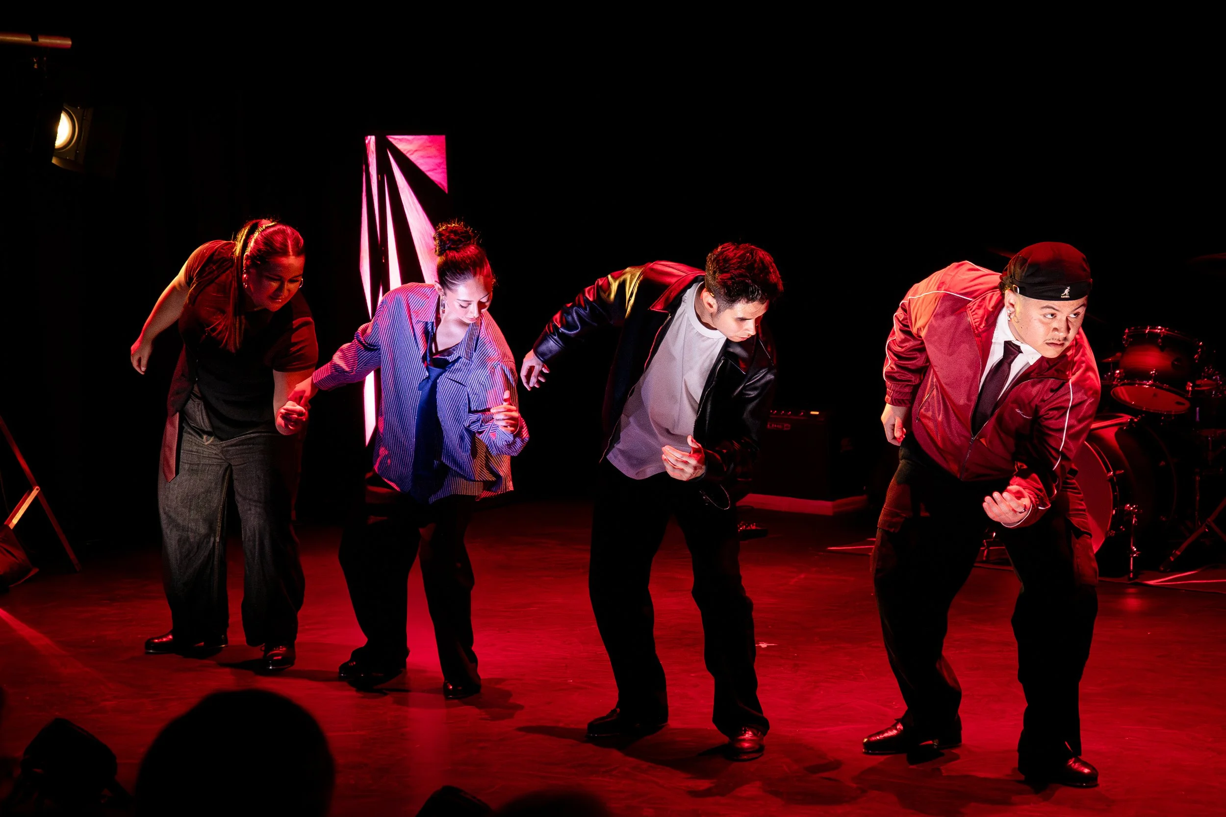 Group of five performers on stage bowing after a performance, illuminated by red stage lighting, with musical instruments in the background.