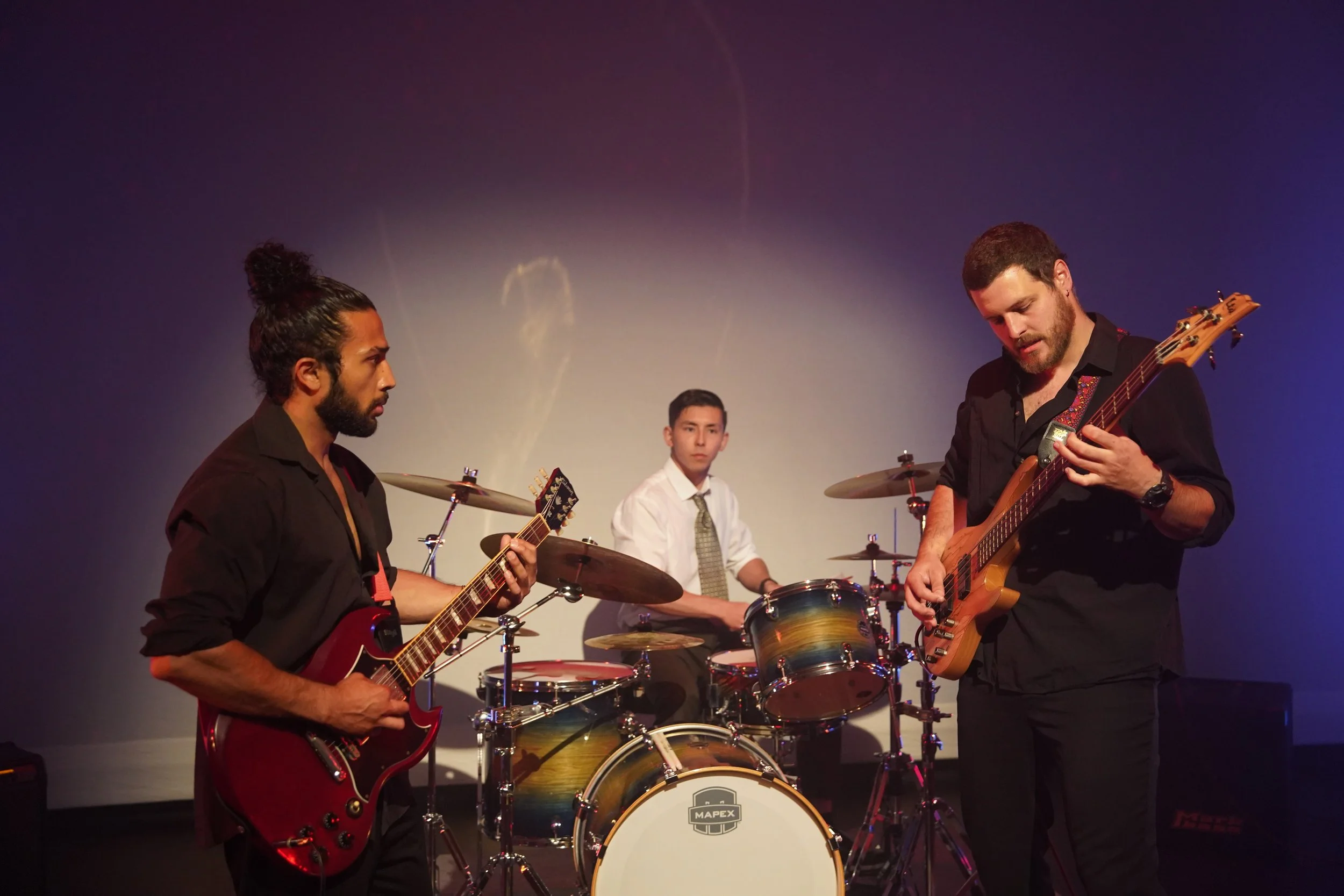 Three musicians performing on stage, playing guitar, drums, and bass guitar, against a purple background.