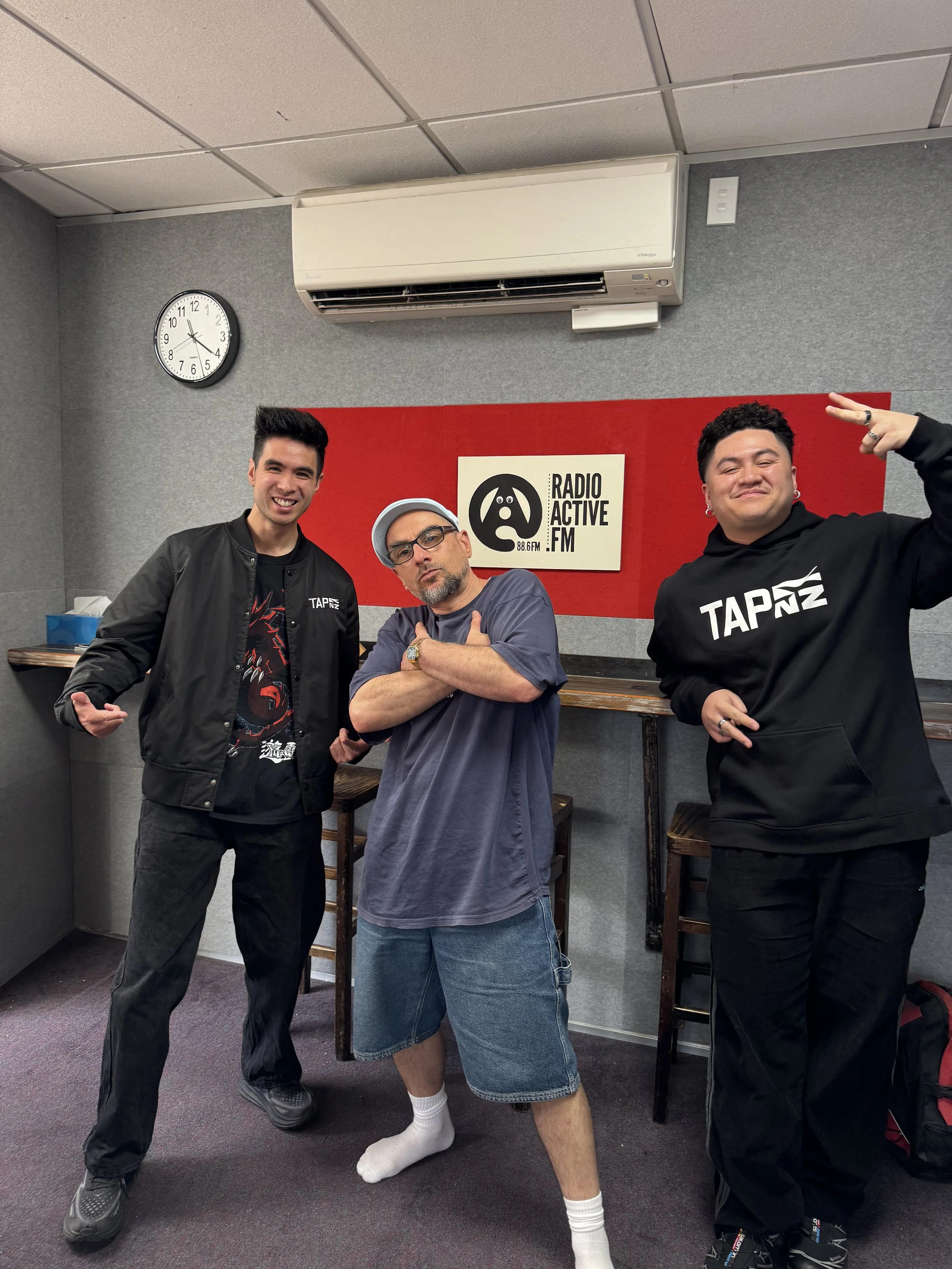 Three men posing in a radio station studio with a red wall and a logo that reads 'Radio Active FM 88.6' in the background. The man in the middle is wearing a casual t-shirt, shorts, and socks, making a gesture with both hands. The man on the left is 