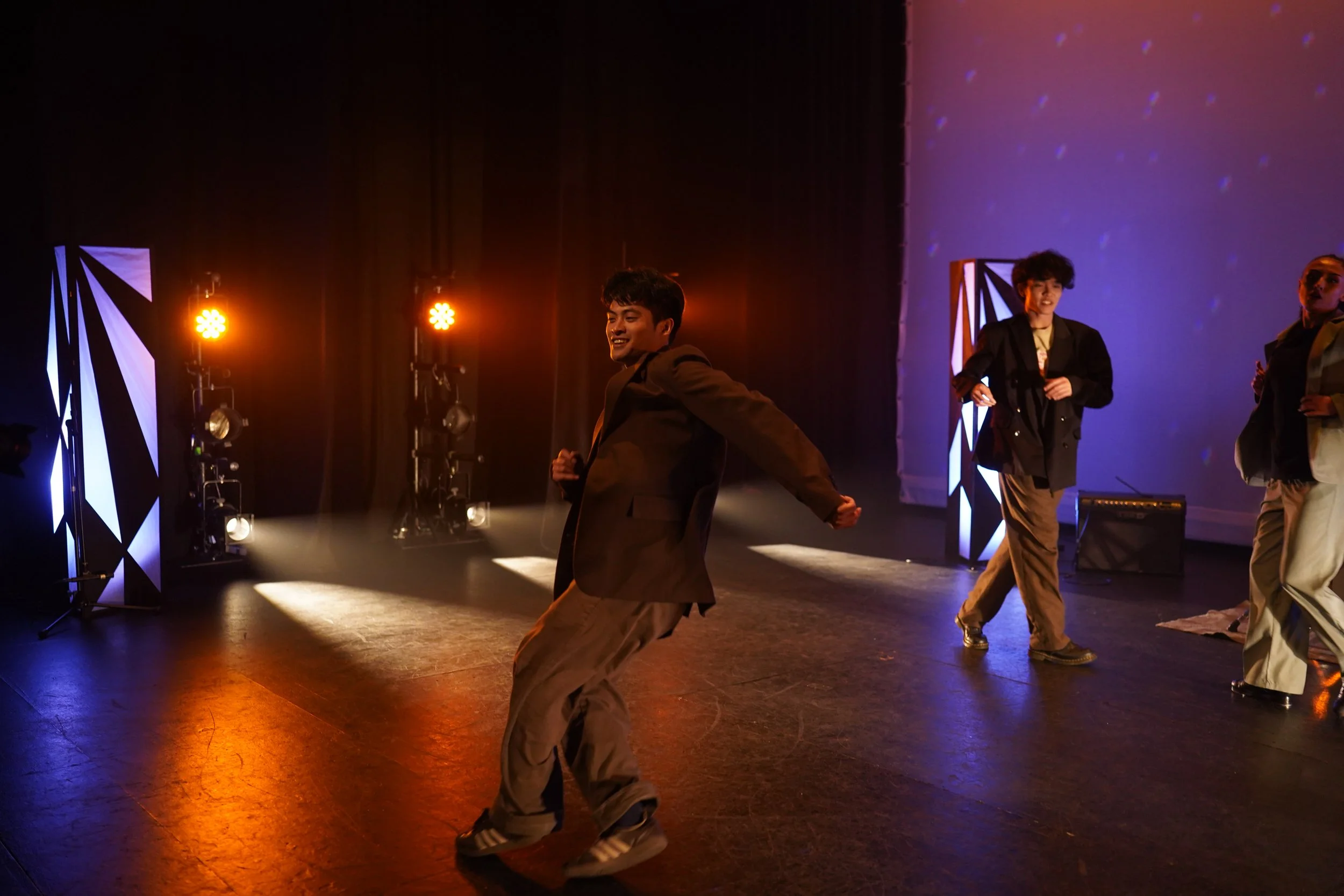 Young men dancing on stage in a performance setting with colorful lighting and abstract background.