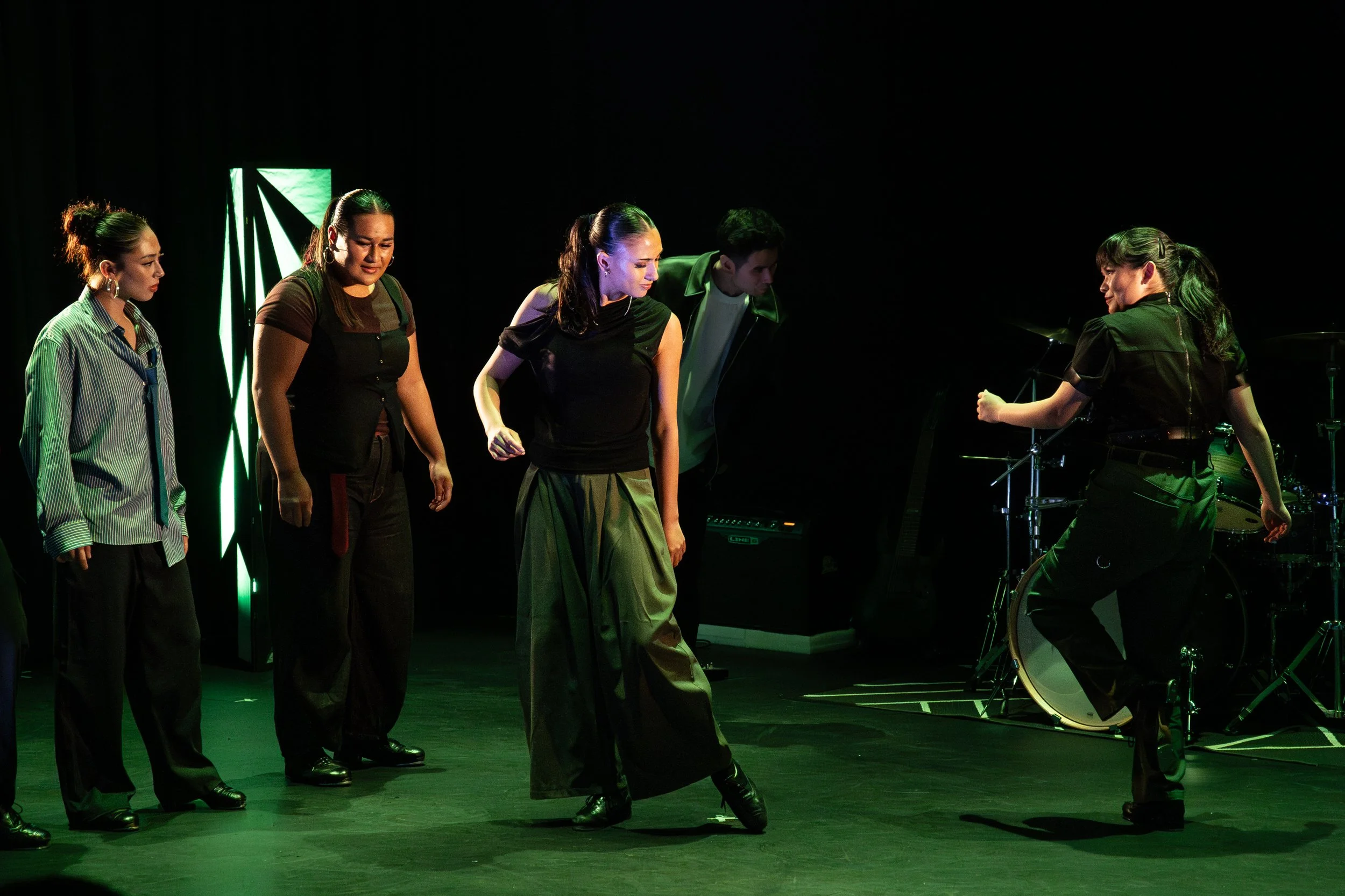 Group of five performers on stage, four women and one man, with musical instruments including drums, dressed in dark clothing, under green stage lighting.