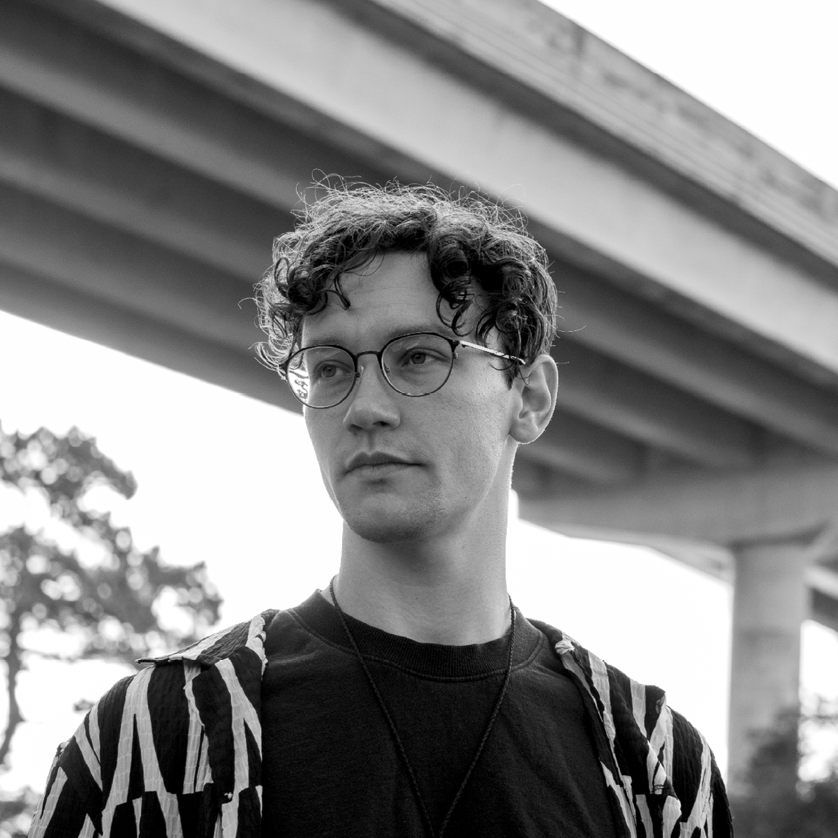 Black and white photo of a young man with curly hair and glasses standing outdoors under a bridge, with trees in the background.