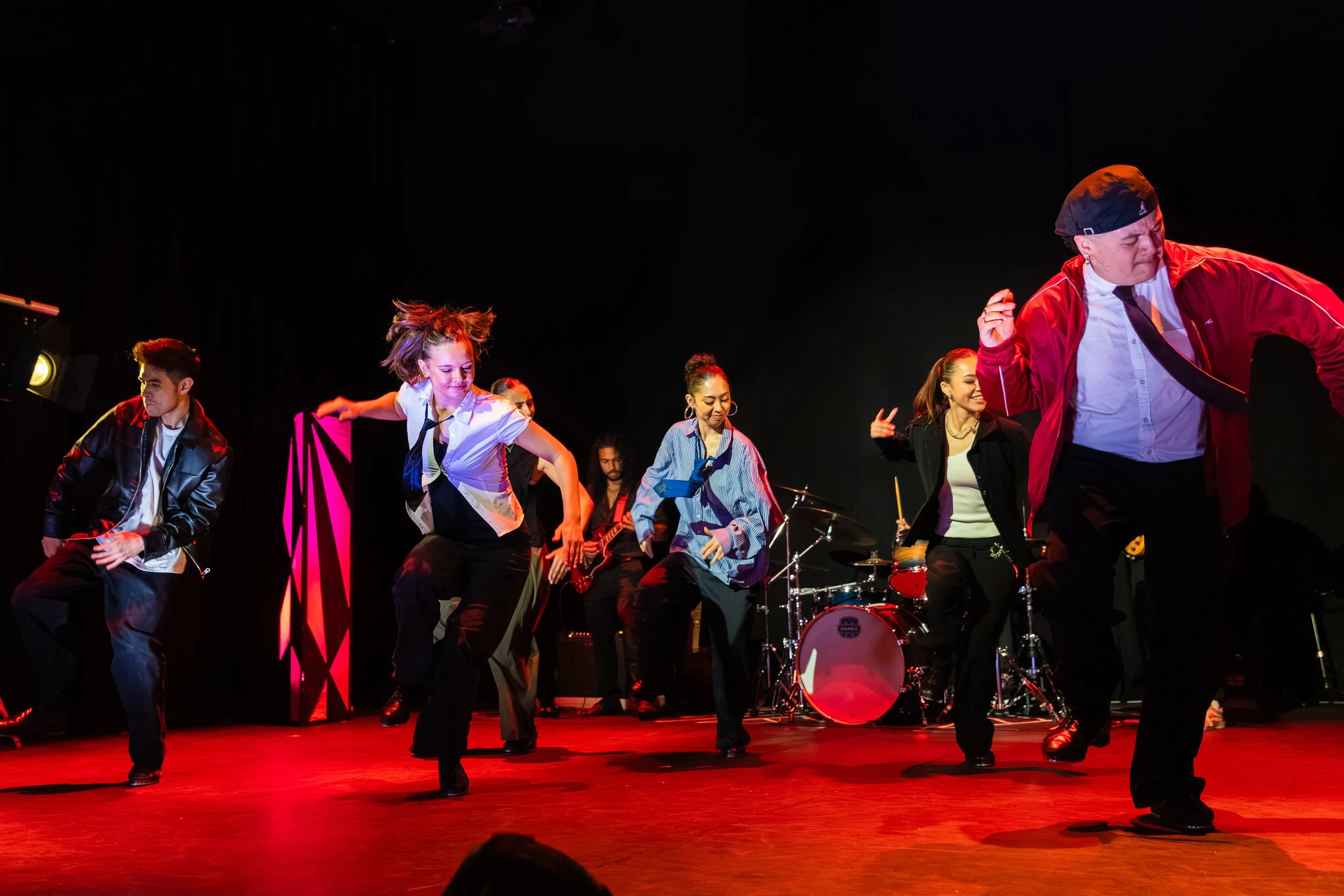 A group of performers dancing on stage with a musical band playing in the background, under red lighting.