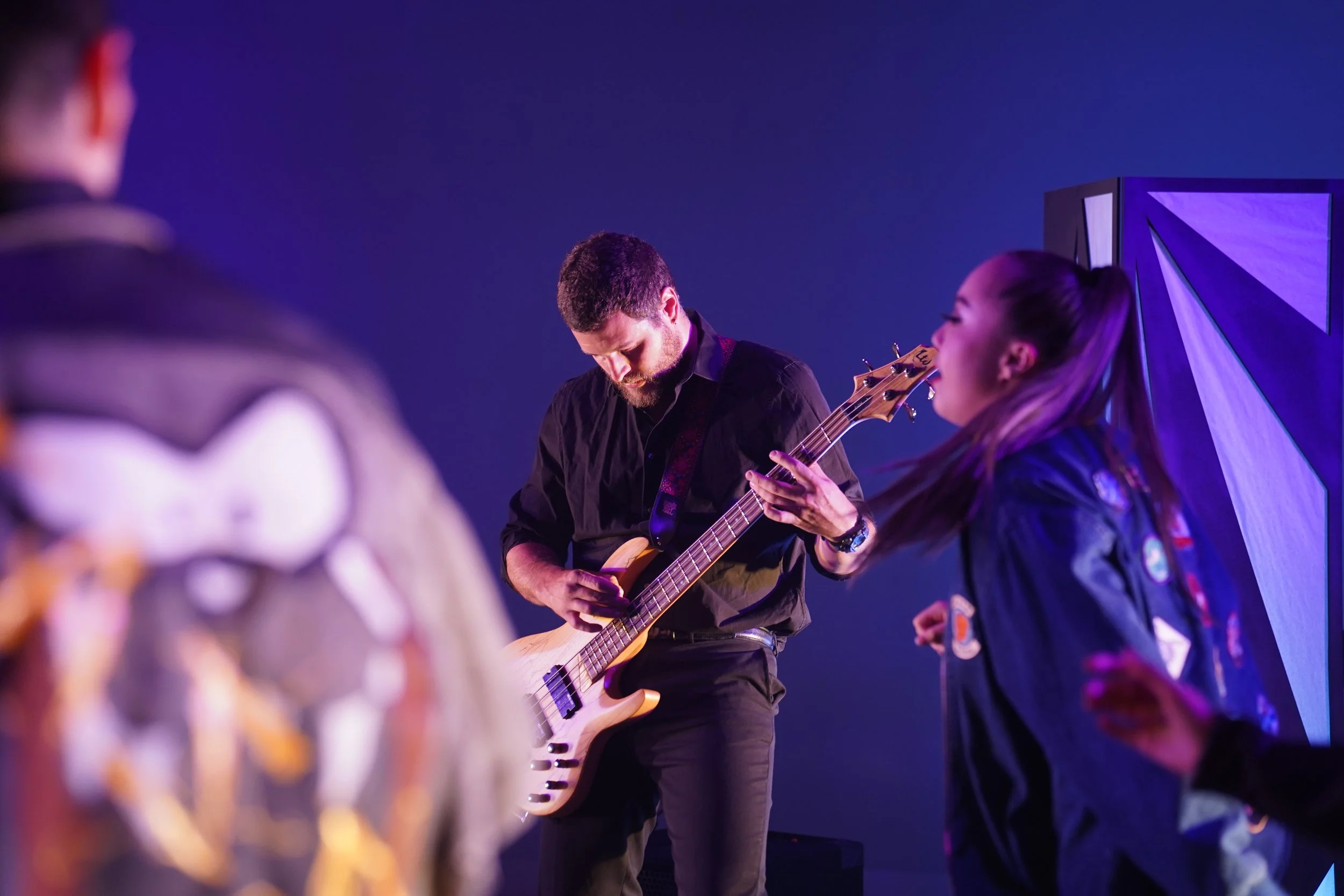 A man playing an electric guitar while surrounded by children in uniforms with patches, in a setting with purple and blue lighting.