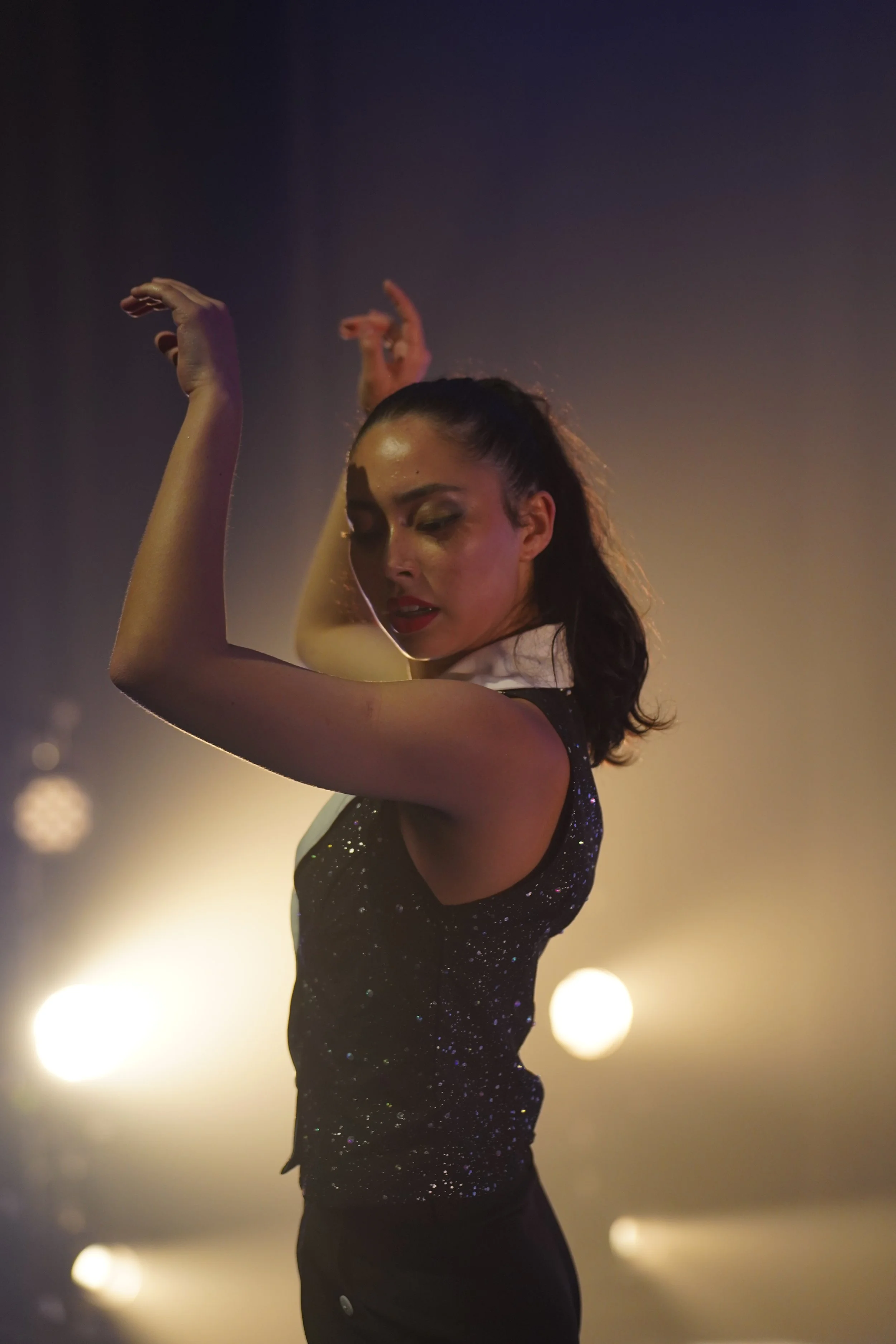 A woman with dark hair in a high ponytail dancing with her eyes closed, wearing a sleeveless, black, glittery top, against a dark background with blurred lights.