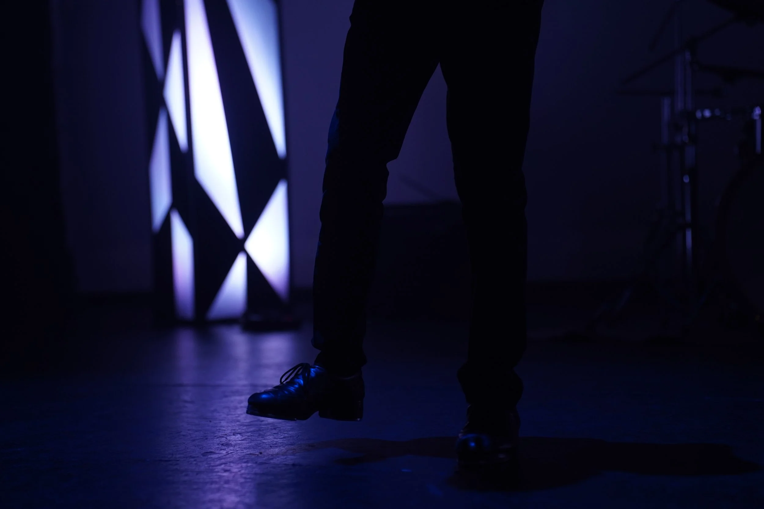 Silhouetted person standing on a stage or studio with blue lighting, with a lit geometric patterned background on the left side.