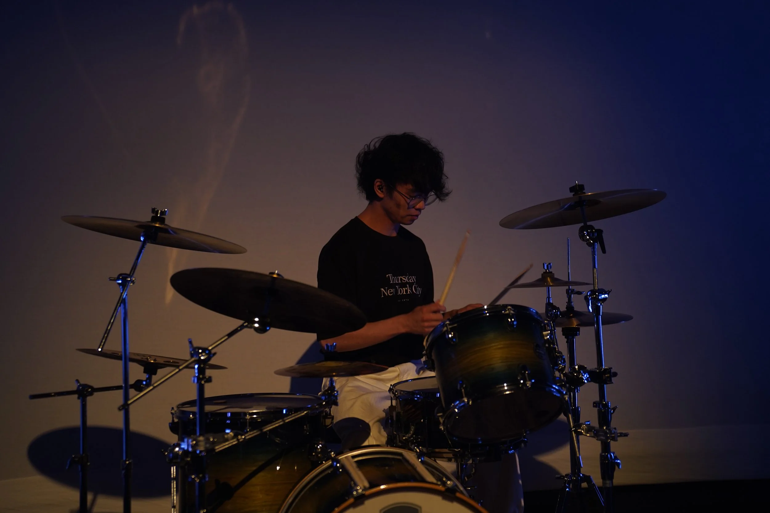 A person with curly hair and glasses playing a drum set in a dimly lit space, wearing a black shirt that says 'Thursday New York City'.