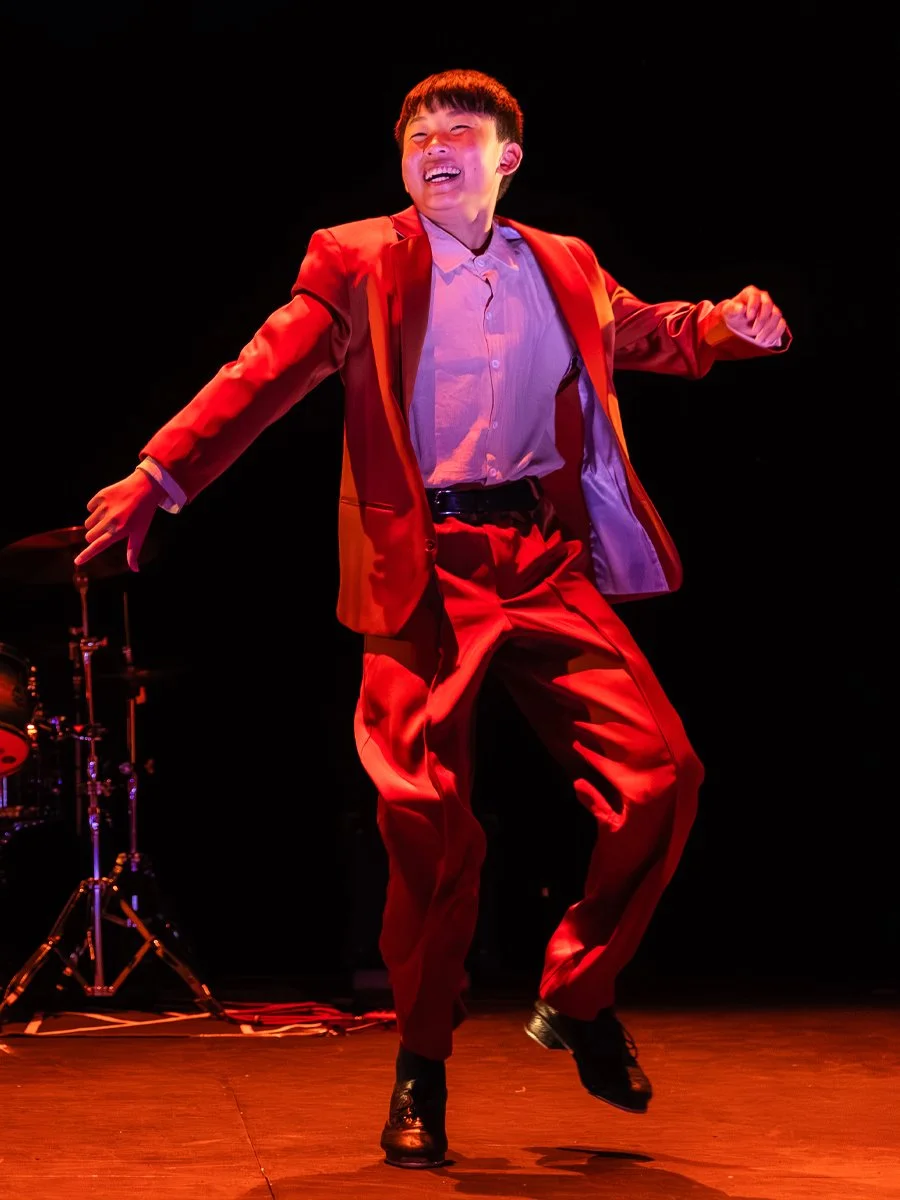 Person dancing on stage, wearing a red suit and black shoes, with a drum set in the background, under stage lighting.