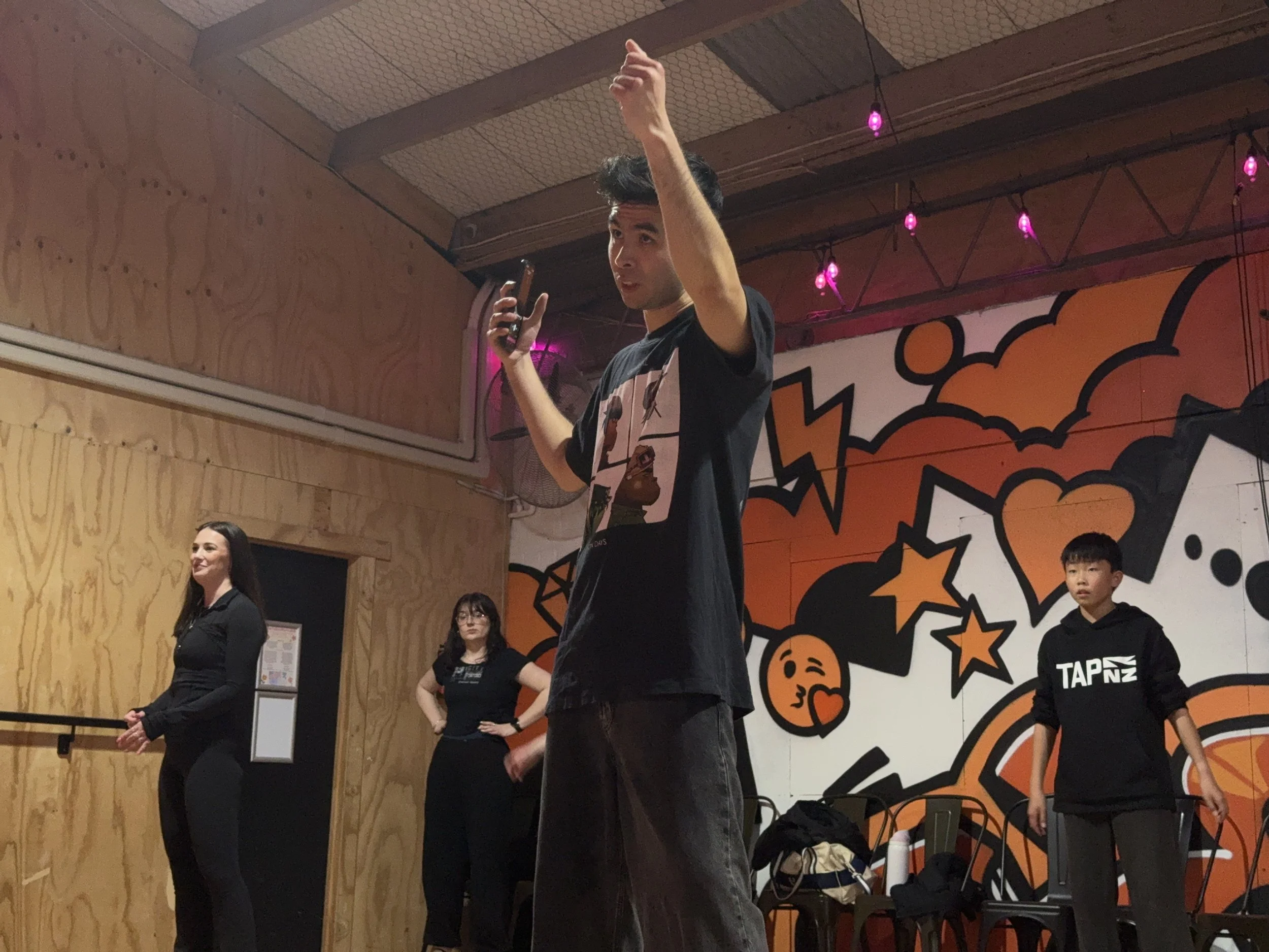 A group of people standing in a room with wood paneling and a wall decorated with orange and black cartoonish images of pumpkins, stars, moons, and bats. One person is raising their hand while holding a phone, and others are standing nearby, some with their arms crossed. The room is decorated for a Halloween event.