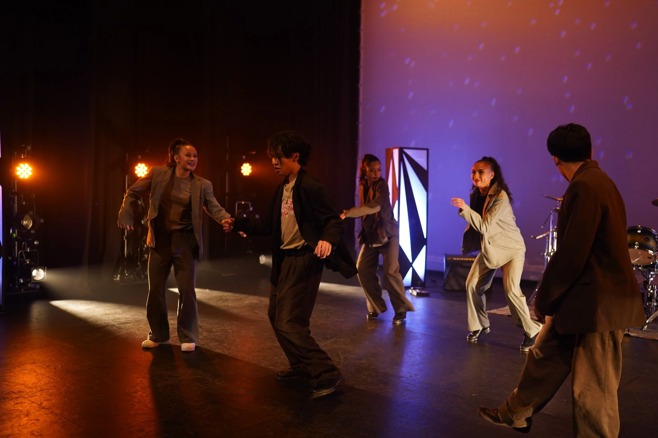 Group of five people dancing on a stage with colorful lights and a backdrop, dressed in casual and semi-formal attire.