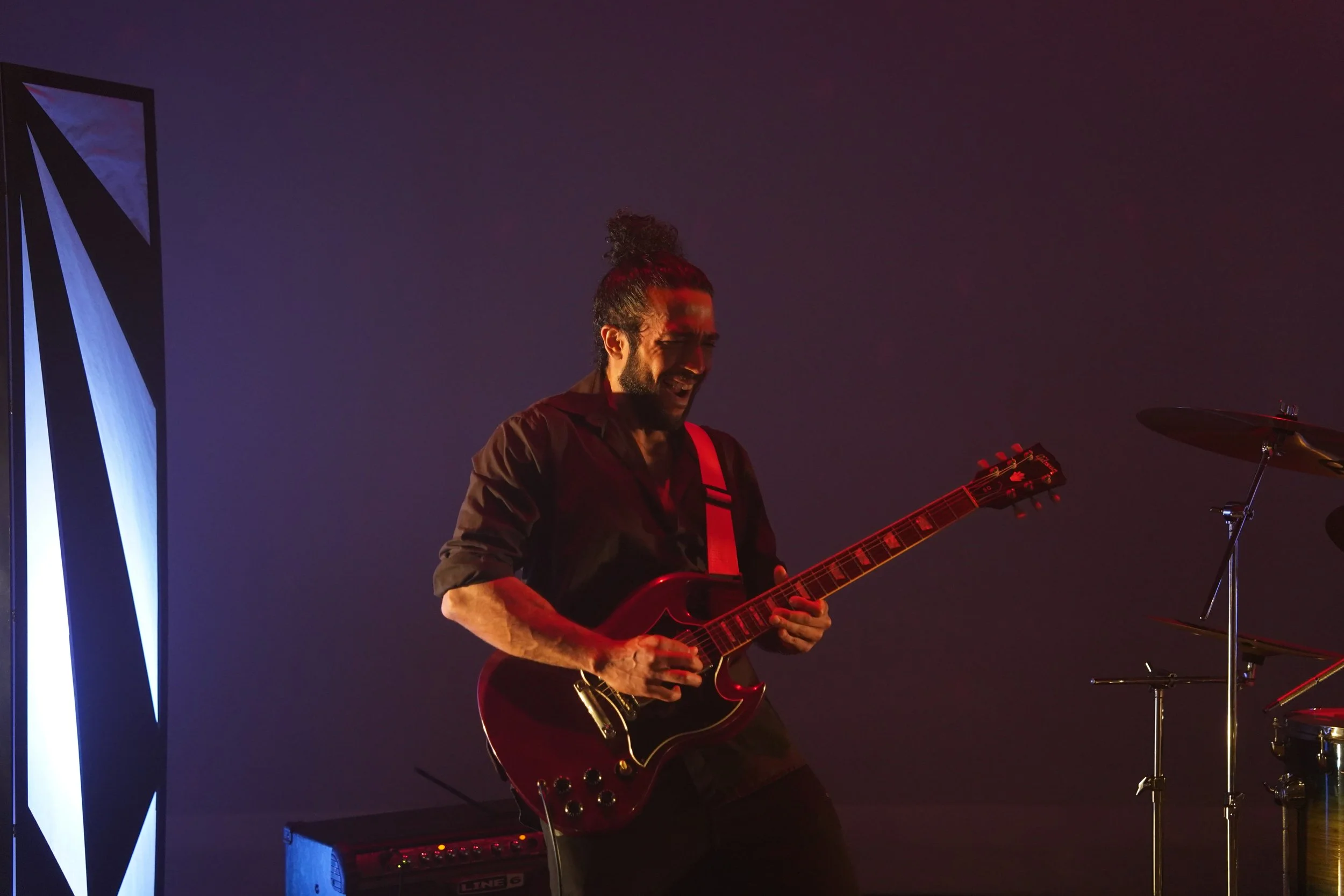 A man with curly hair tied up in a bun is playing an electric guitar on stage, smiling and enjoying the moment, with a drum set and electronic equipment around him, against a dark and colorful background.