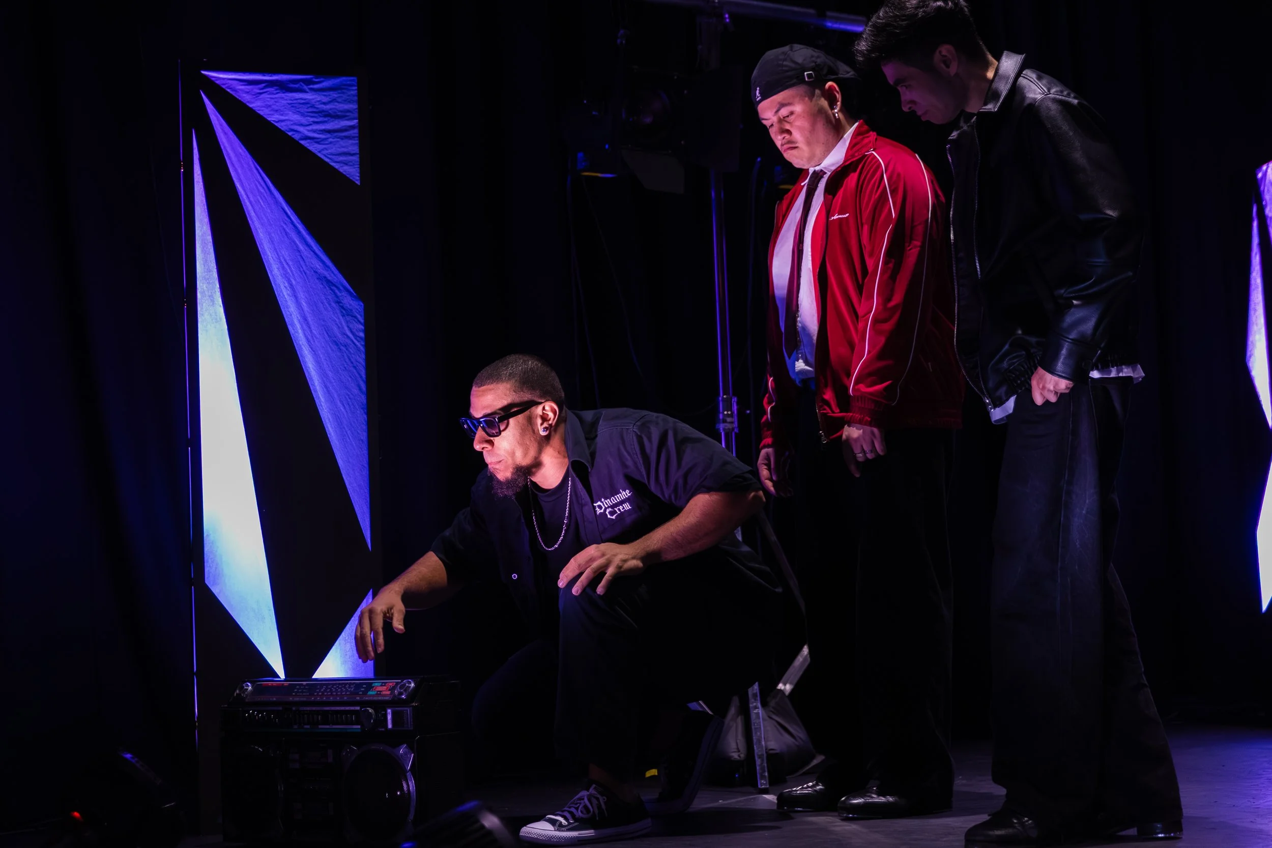 Three young men are on a dark stage, illuminated by purple and blue stage lights. One man is sitting, wearing glasses and a black shirt, working with electronic music equipment. Two men are standing; one wearing a red jacket and black cap, the other in a black jacket, both watching the seated man work.