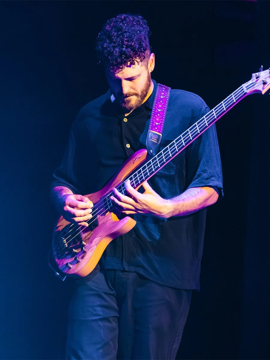 A musician with curly hair and a beard playing an electric bass guitar on stage, illuminated by purple and blue stage lights.