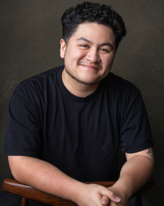 Portrait of a young man with dark, curly hair, smiling, wearing a black t-shirt, sitting with arms crossed on a wooden chair against a dark background.
