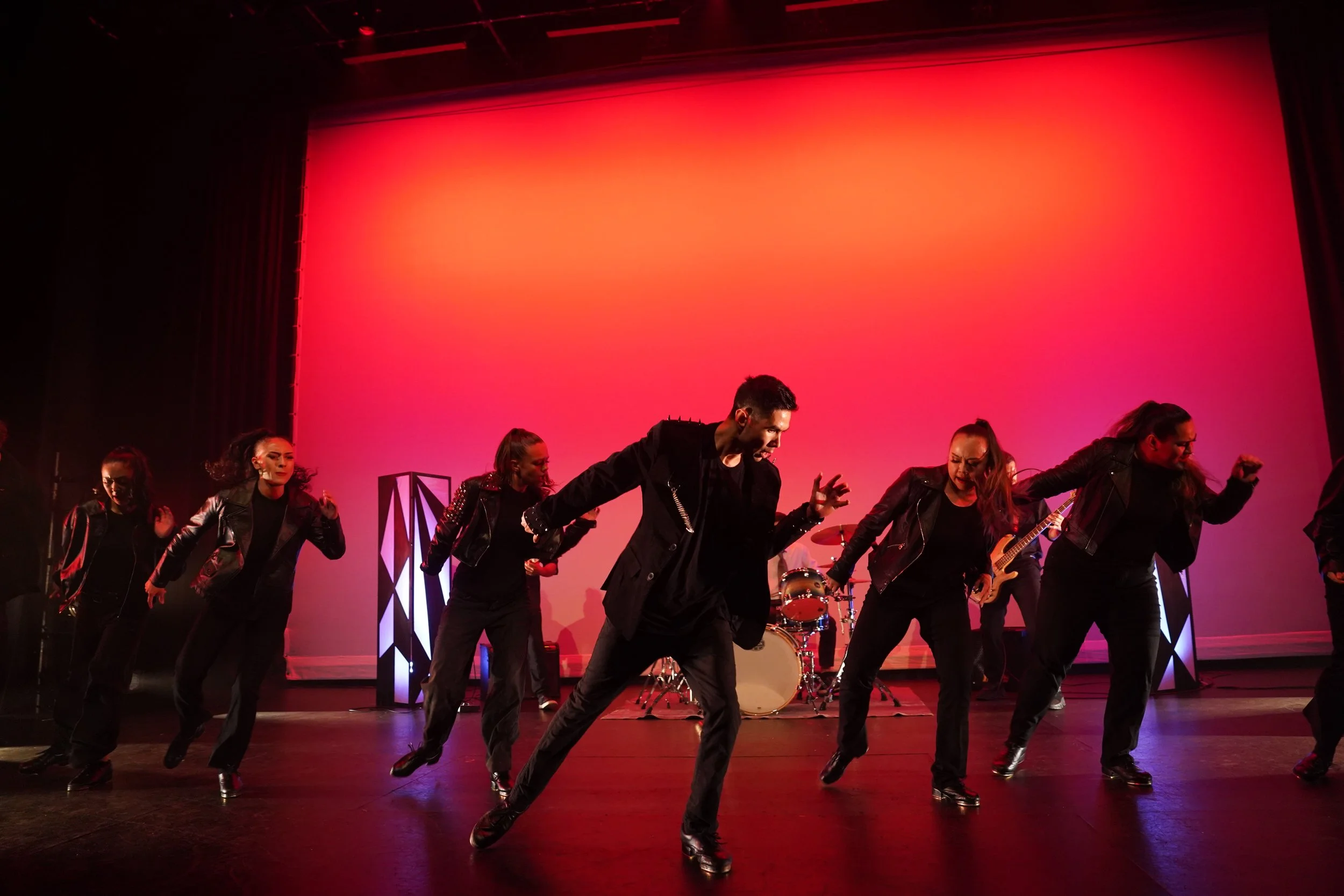 A group of dancers performing on stage with a vibrant pink and orange backdrop, dressed in black clothing, with some playing instruments.