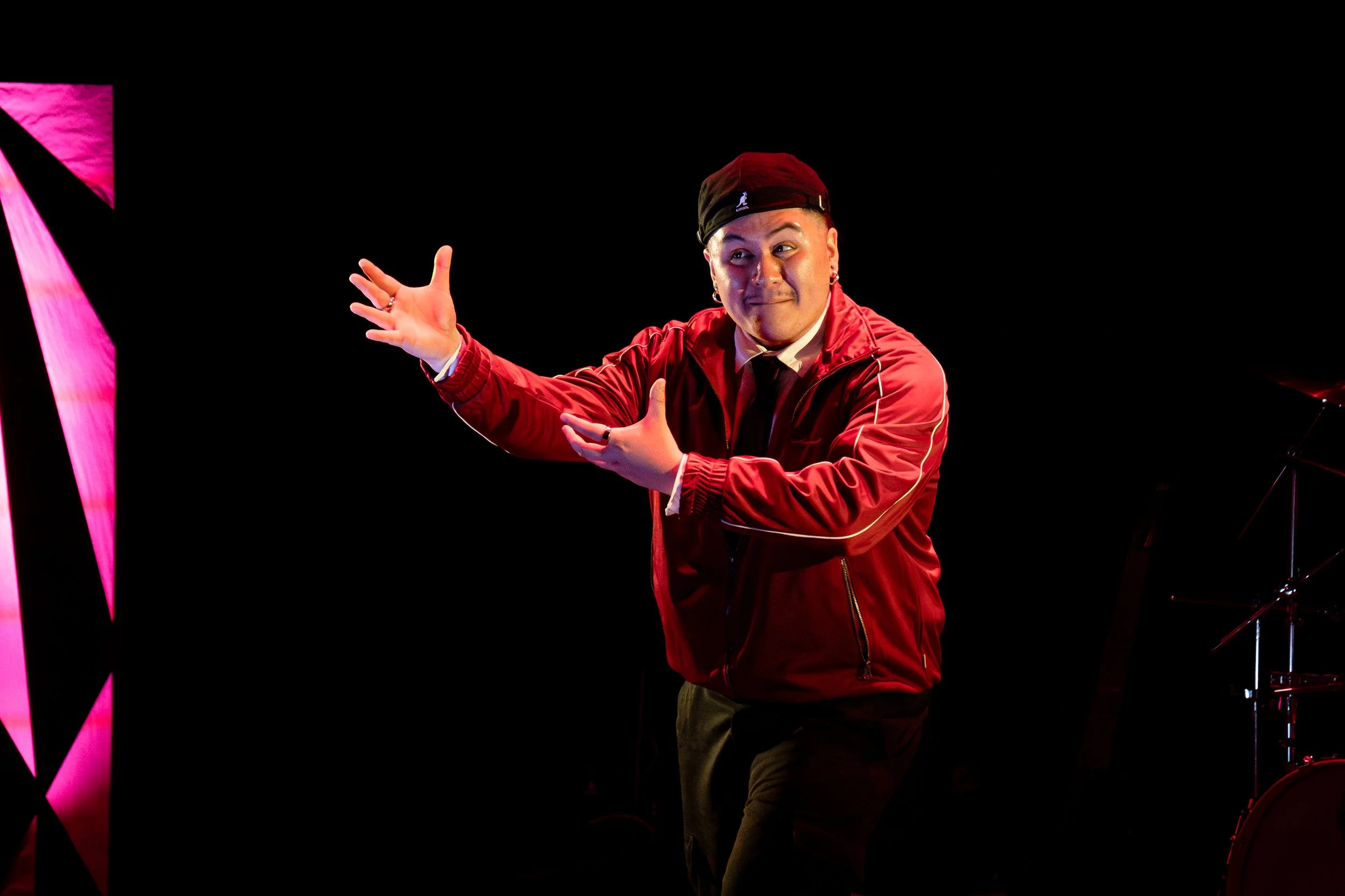A man wearing a red jacket and black hat performing on stage, with a pink geometric structure partially visible on the left.