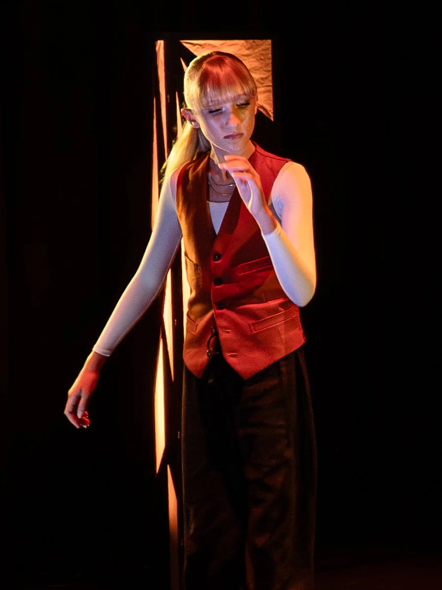 A woman with blonde hair in a ponytail, wearing a red vest, white long-sleeve shirt, and black pants, standing on stage with a dark background and some lighting.