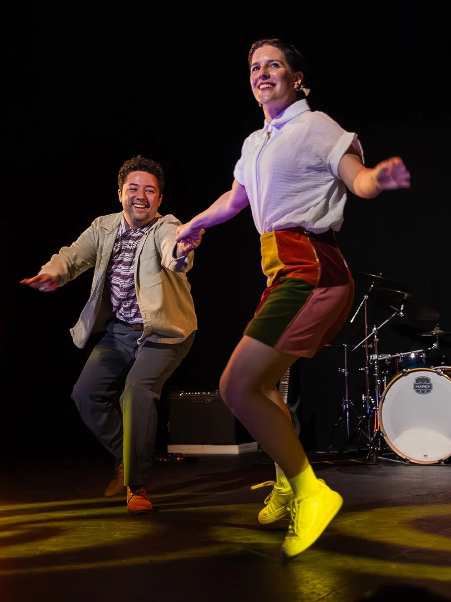 A man and woman dancing on a stage, smiling and holding hands. The woman is wearing a white shirt, colorful shorts, and yellow shoes. The man is dressed in a beige jacket, striped shirt, and dark pants. There is musical equipment, including drums, in the background.