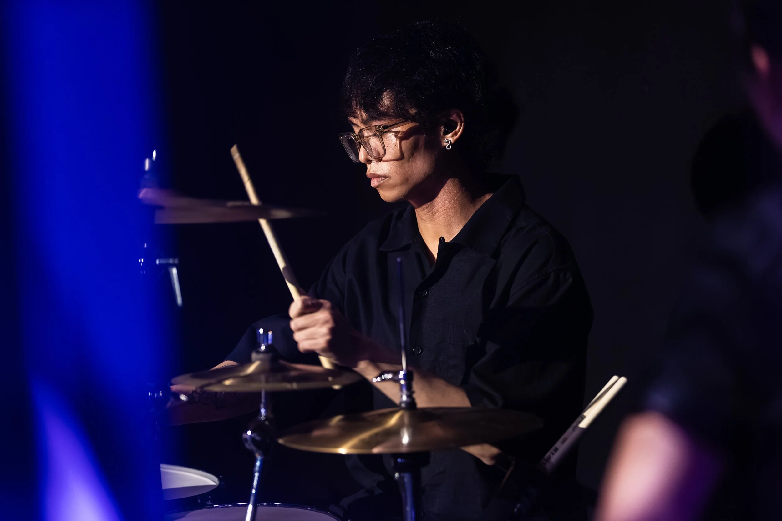 A musician wearing glasses and earrings playing drums on stage with dark background, illuminated by stage lights.