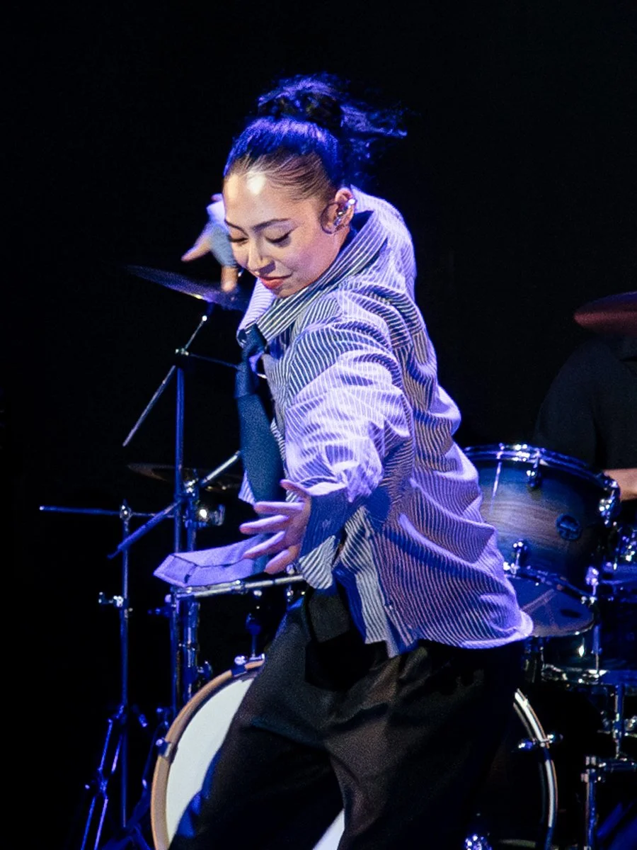 A woman dancing or performing on stage with a drummer behind her, illuminated by stage lights, wearing a striped shirt and a loose tie.