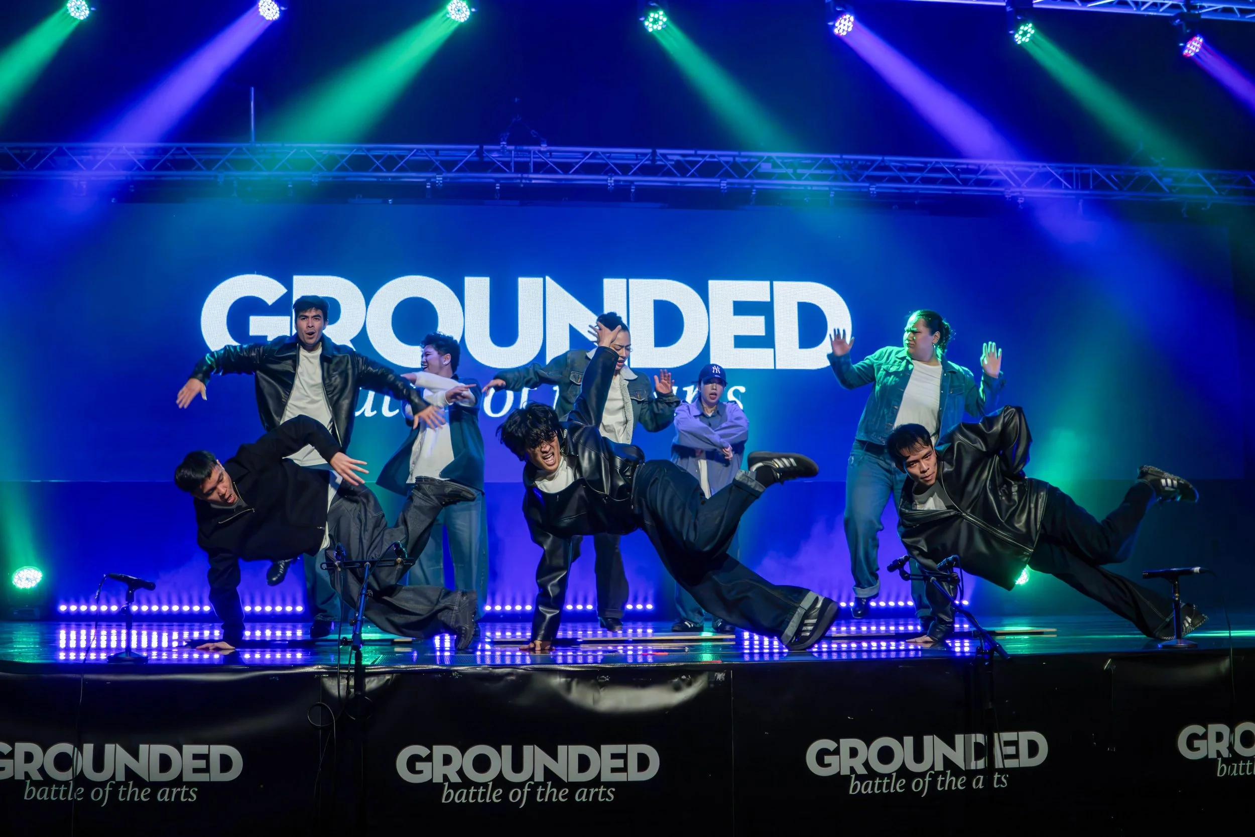 A group of dancers performing on stage under colorful lighting with a large screen in the background displaying the word 'GROUNDED' and the phrase 'battle of the arts'.
