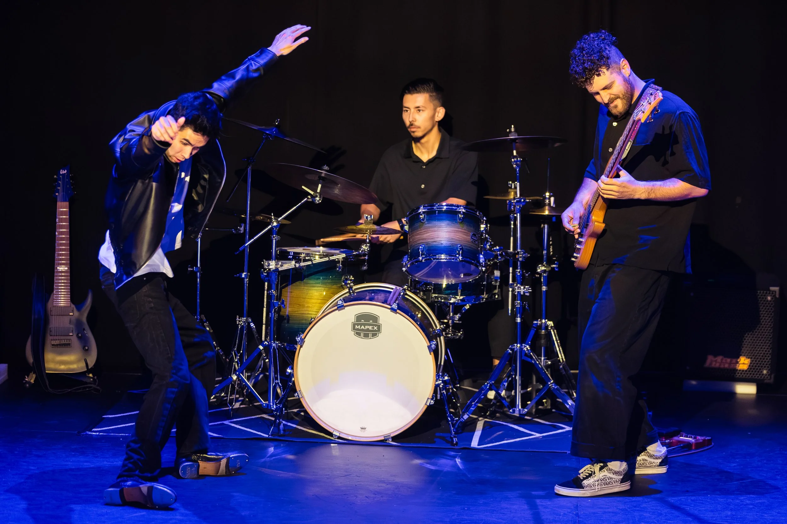 Three musicians performing on stage with black background, two playing guitars and one on drums, illuminated by stage lights.