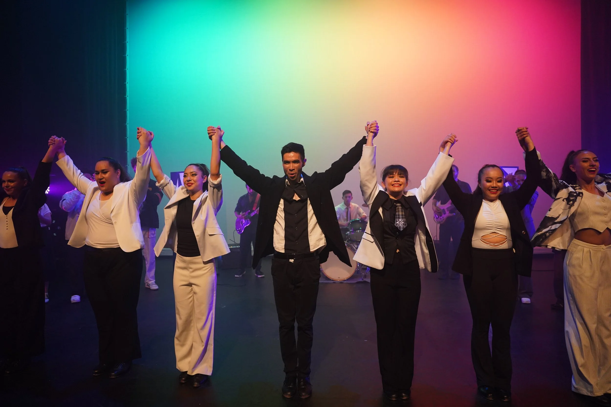 Group of dancers holding hands with arms raised, performing on stage with colorful lighting and live band in the background.