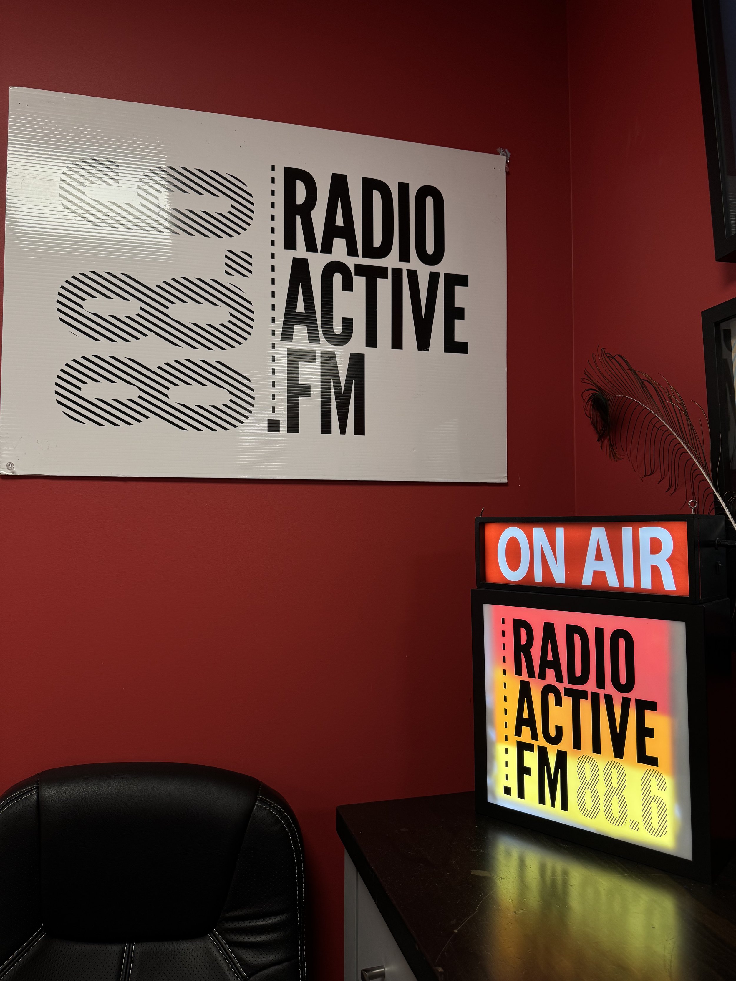 Radio Active FM 99.6 signage on a illuminated light box in a recording or radio studio, with a red wall background and a black chair nearby.