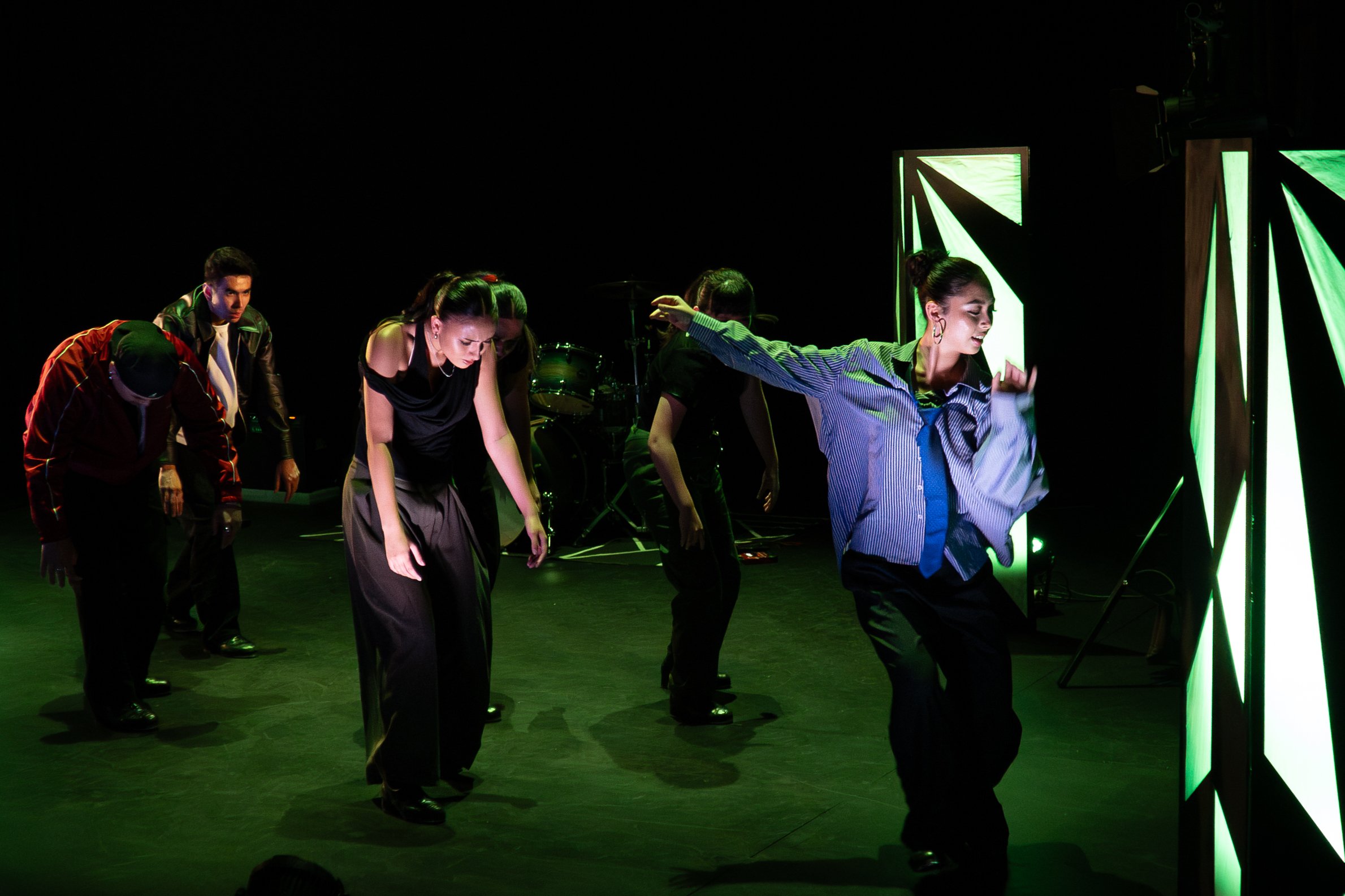 Group of dancers performing on stage with green lighting and abstract set pieces, dressed in dark and casual clothing.