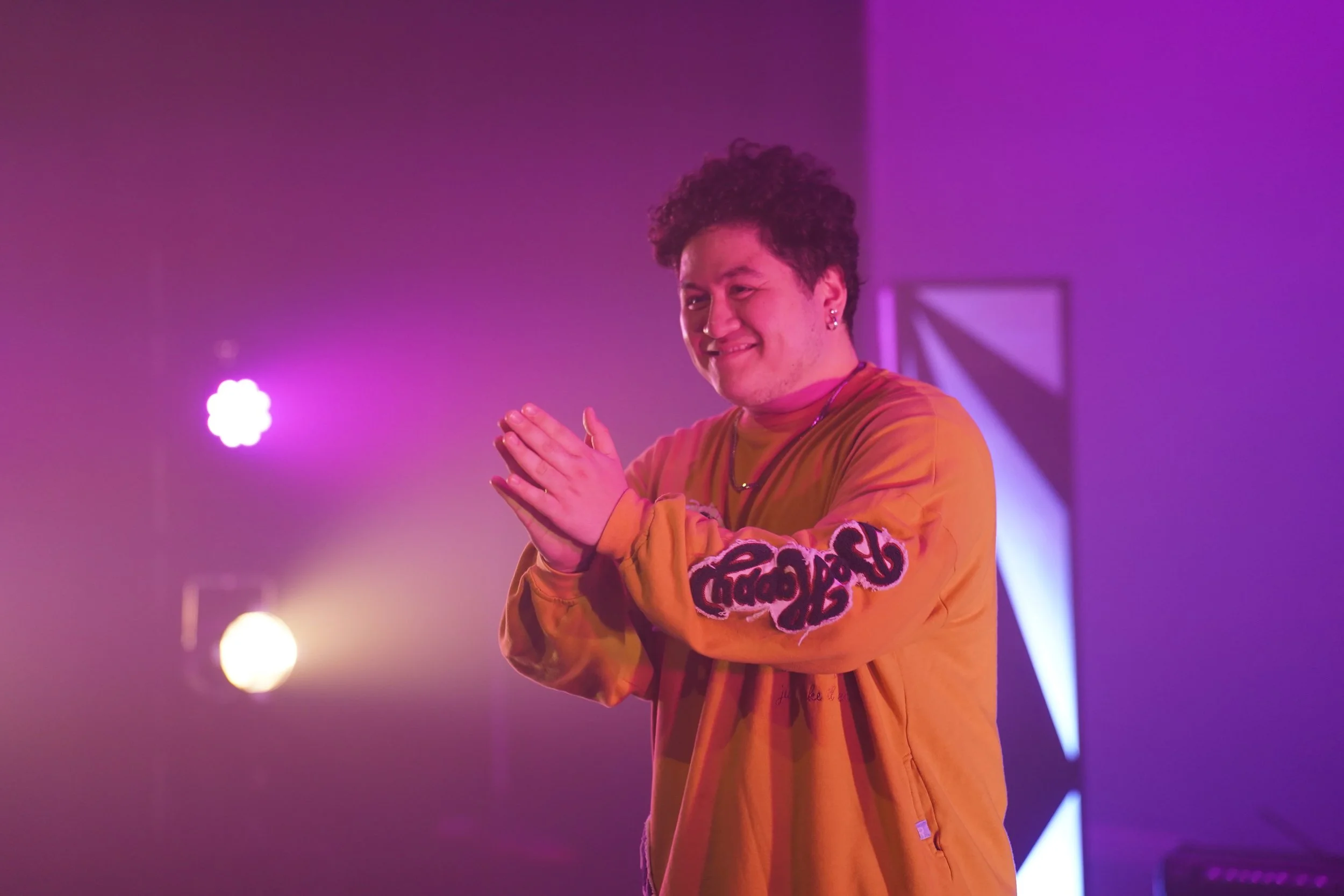 A young man in an orange jacket with a smile, making a hand gesture against a colorful purple and pink lit background.
