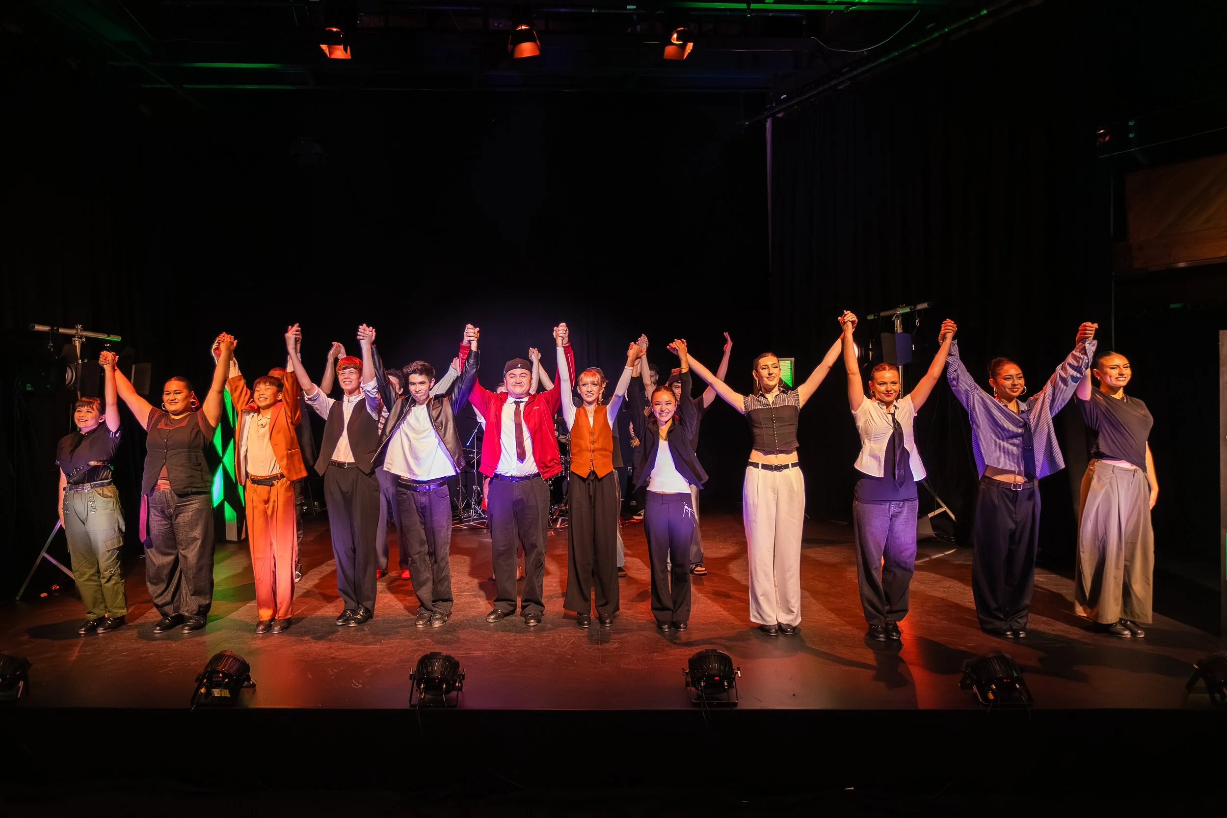 A group of people on stage holding hands and taking a bow after a performance, with stage lights and equipment visible.