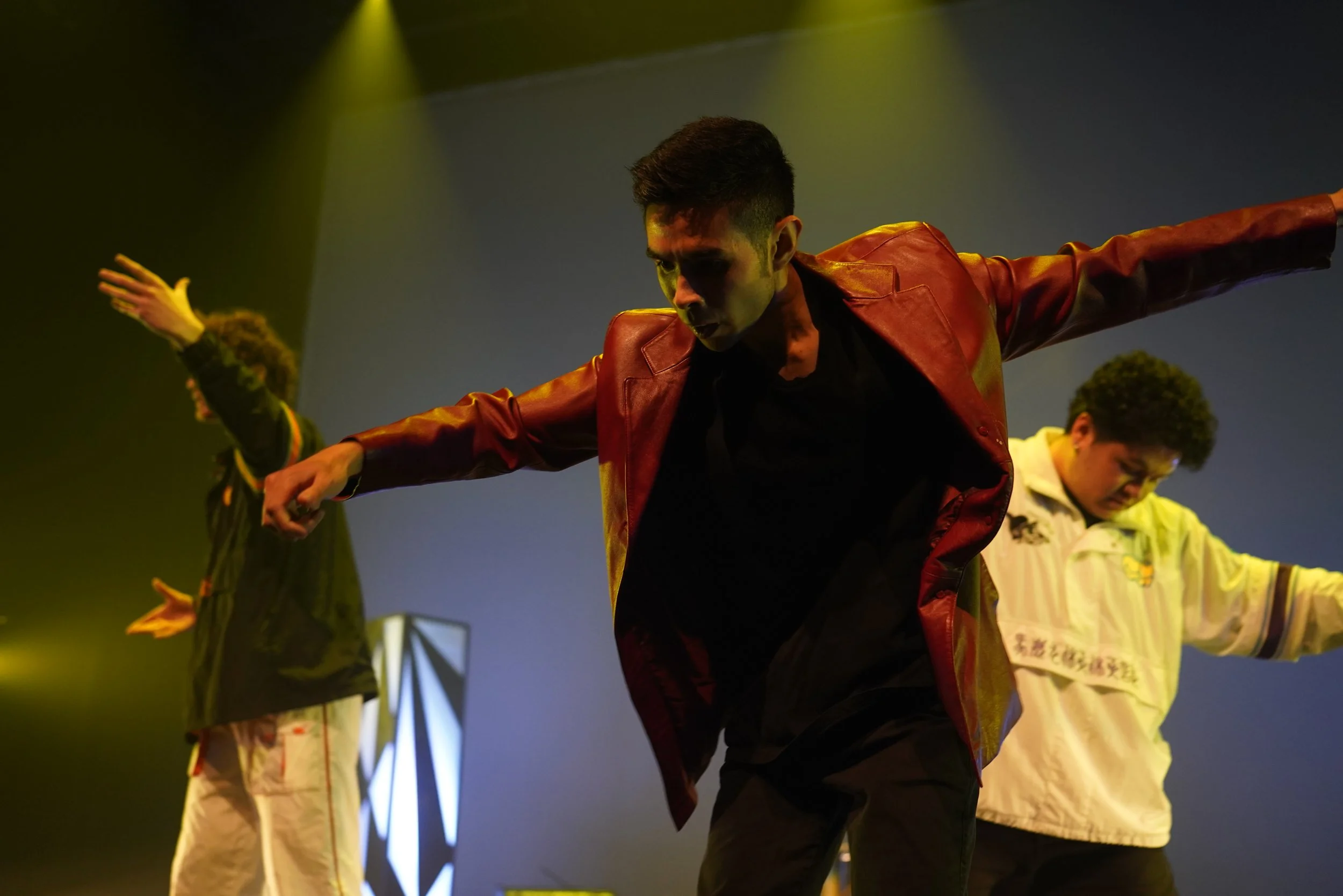 Performer with arms outstretched on stage, wearing a red leather jacket, with other performers in the background under stage lighting.