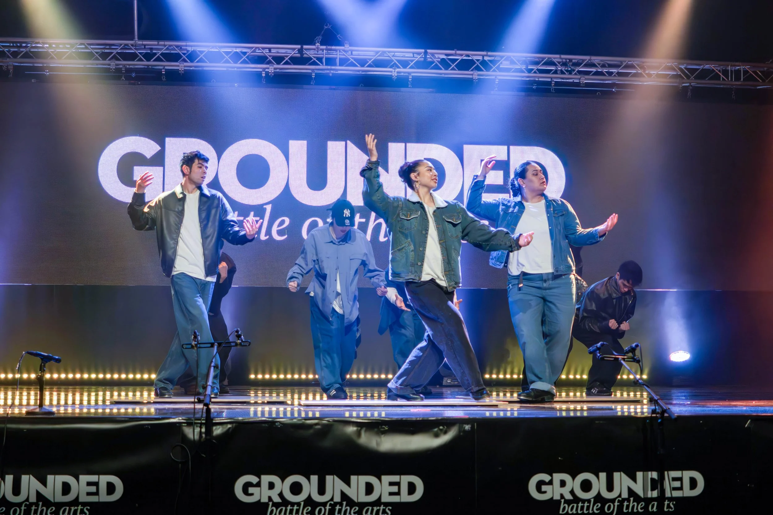 Group of dancers performing on stage with a large screen behind them displaying the word 'GROUNDED'. The dancers are dressed casually and are in various dance poses. Stage lighting emphasizes the performance.