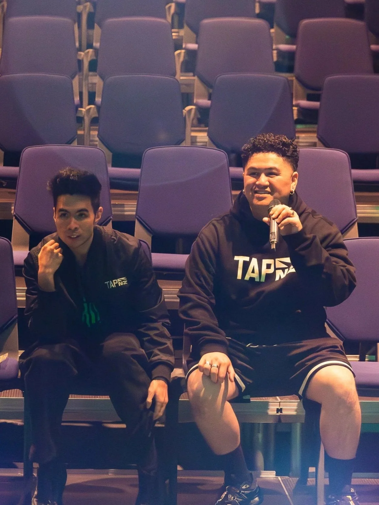 Two young men sitting in stadium seats, one holding a microphone and wearing a black hoodie with 'TAP' logo, the other making a gesture near his face, both dressed in black with matching logos, surrounded by empty purple stadium seats.
