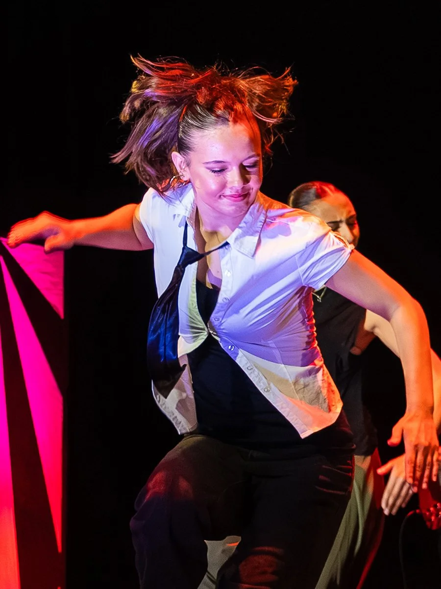 A woman with short, tousled hair dancing on stage, wearing a white shirt and black pants, with colorful stage lighting.