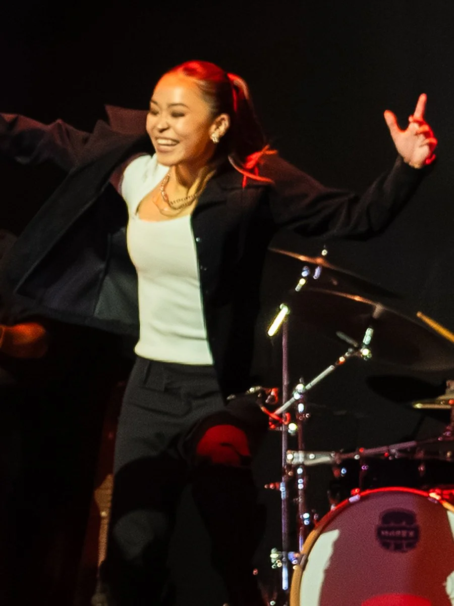 A woman with long hair tied back, wearing earrings, a white top, and a black jacket, is smiling and dancing near a drum set on a dark stage.